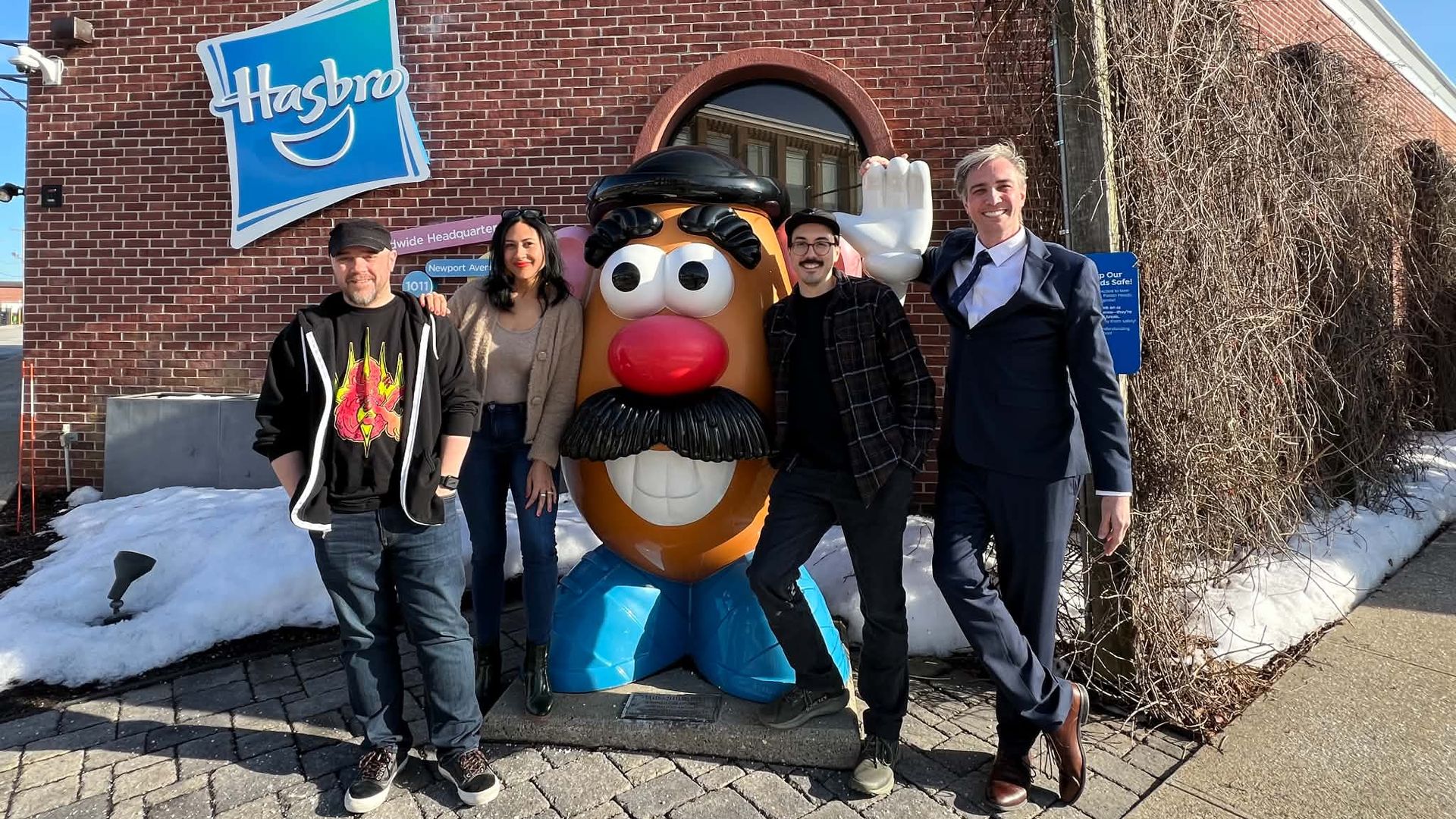 Five people stand in front of a red brick Hasbro building with a large Mr. Potato Head statue. Clear blue sky, patches of snow, and bare vines along the wall.
