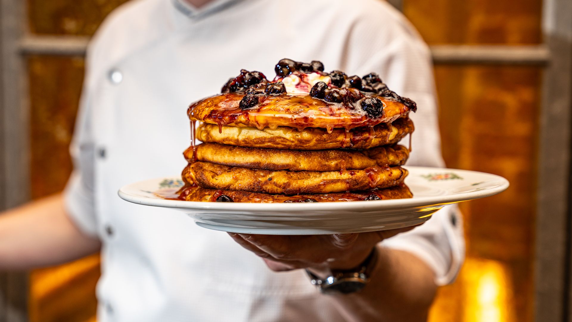 Stack of four golden pancakes topped with blueberries, melted butter, and dripping syrup, held on a white plate by a person in a white chef coat with a warm, orange background.