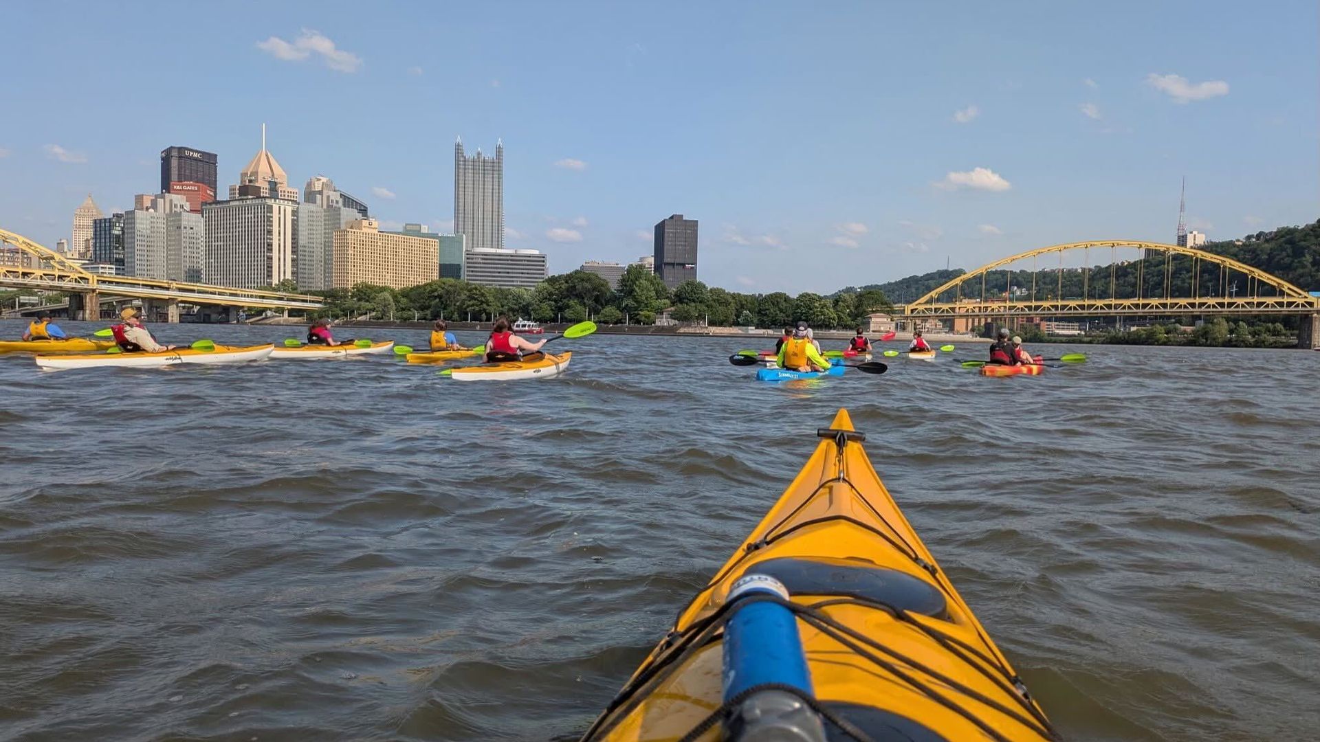 View from a yellow kayak on a river with multiple kayakers ahead in colorful kayaks; a city skyline with tall buildings and a yellow arched bridge under a clear blue sky in Downtown Pittsburgh