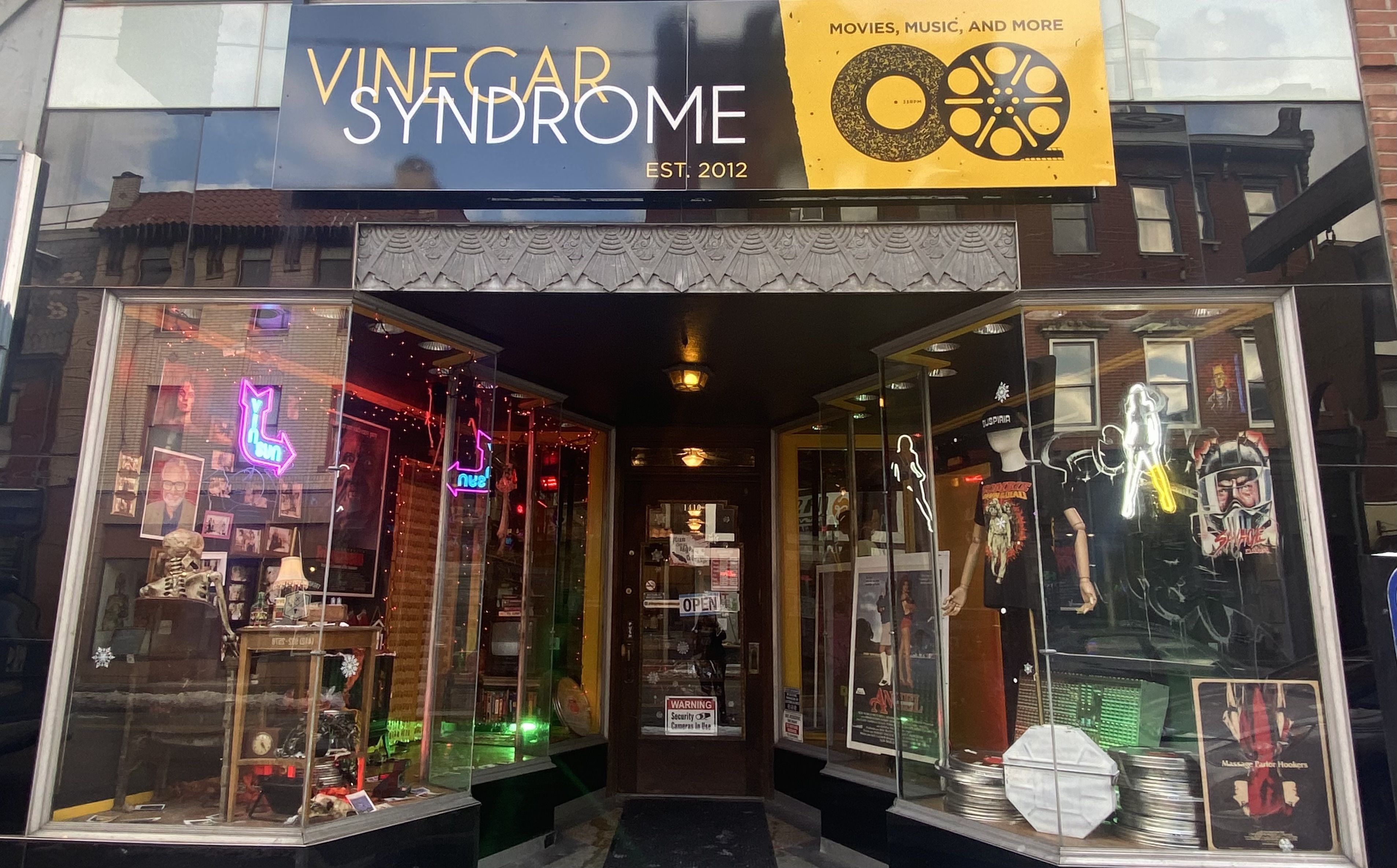 Storefront of Vinegar Syndrome, a movie-focused shop with skeleton decor, neon signs, movie posters, and vintage film reels displayed in glass windows.