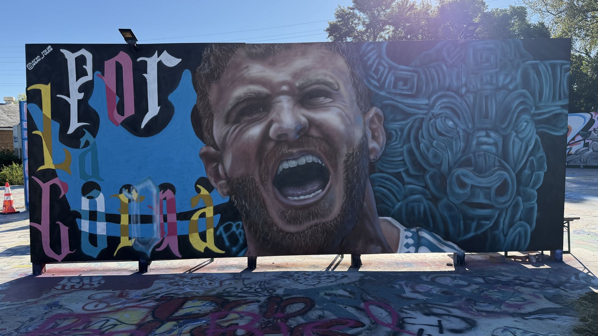 Outdoor mural on a black wall features a screaming man's face at center, with colorful Gothic letters on the left and blue swirling shapes to the right; graffiti on the ground in front.