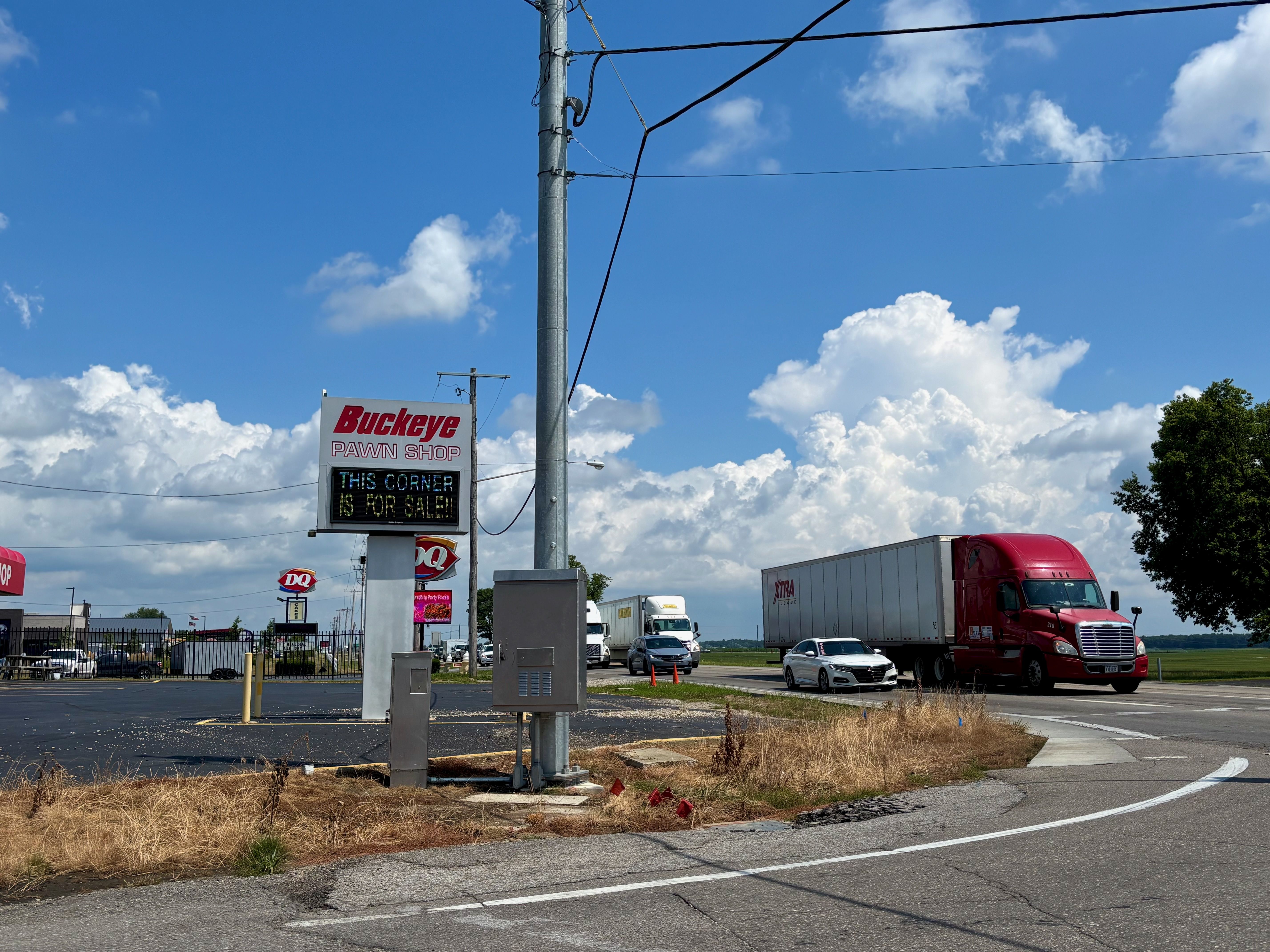 Clear blue sky with white clouds. A tall gray utility pole with wires rises above a Buckeye Pawn Shop sign reading "This corner is FOR SALE!"; a red semi-truck and parked cars fill the lot.