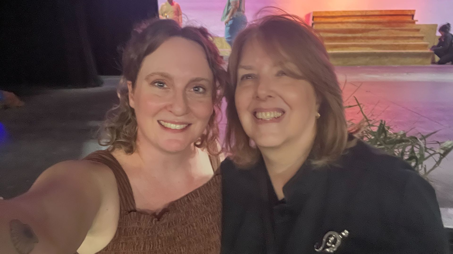 Two women smile for a selfie on a stage with pink lighting. Left wears a brown sleeveless top with a tattoo on her arm; right wears a black jacket with a pin. Steps and people on stage in background.