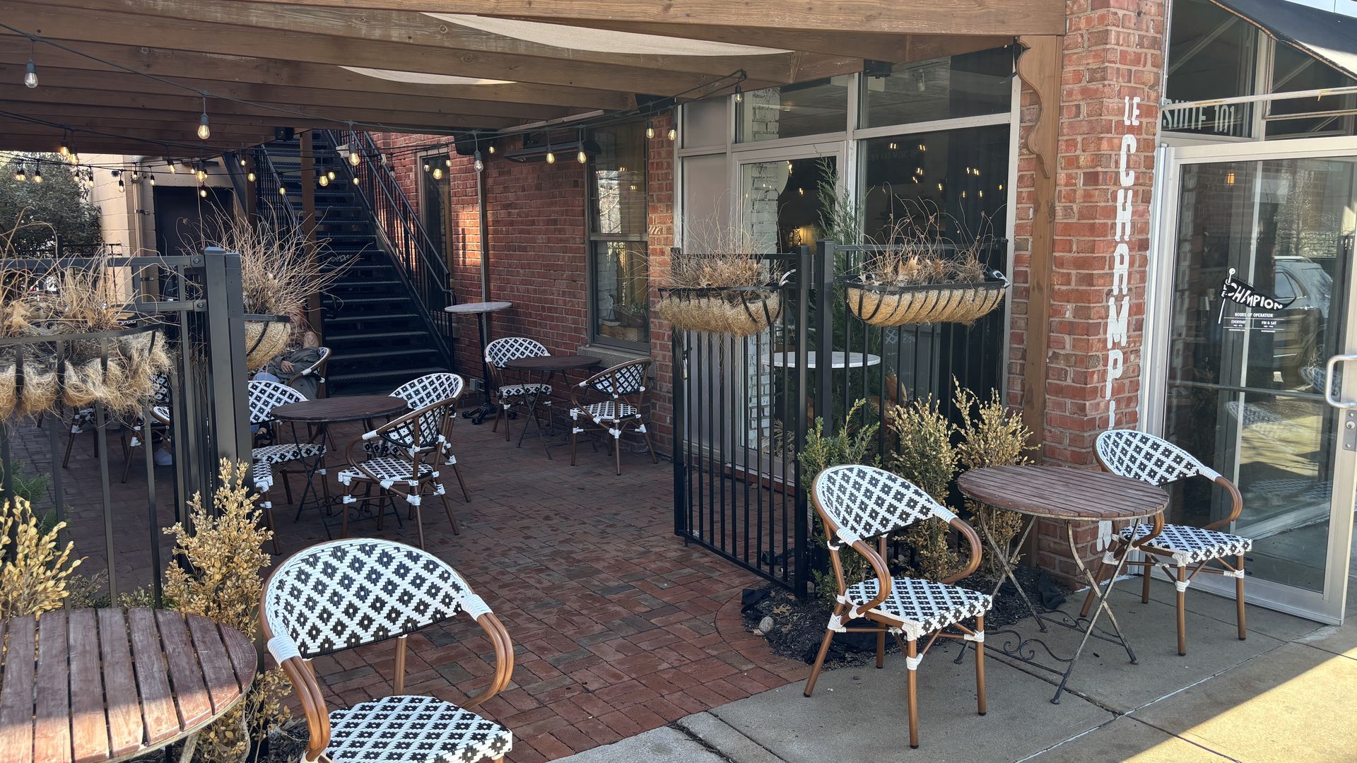 Outdoor cafe patio with string lights under a wooden pergola, brick building, black metal railings, round tables, and white-and-black patterned chairs; planters with dried grasses.