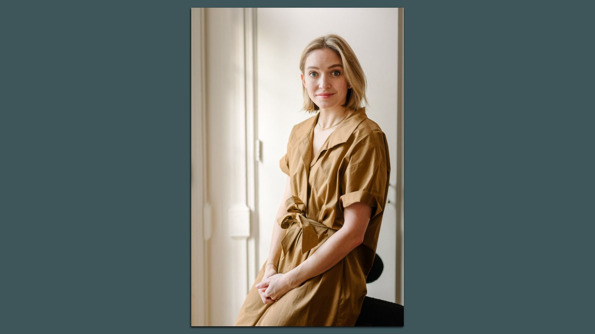 Blonde woman in a light brown dress with rolled-up sleeves stands near a white wall, looking at the camera with a slight smile, hands gently clasped in front.