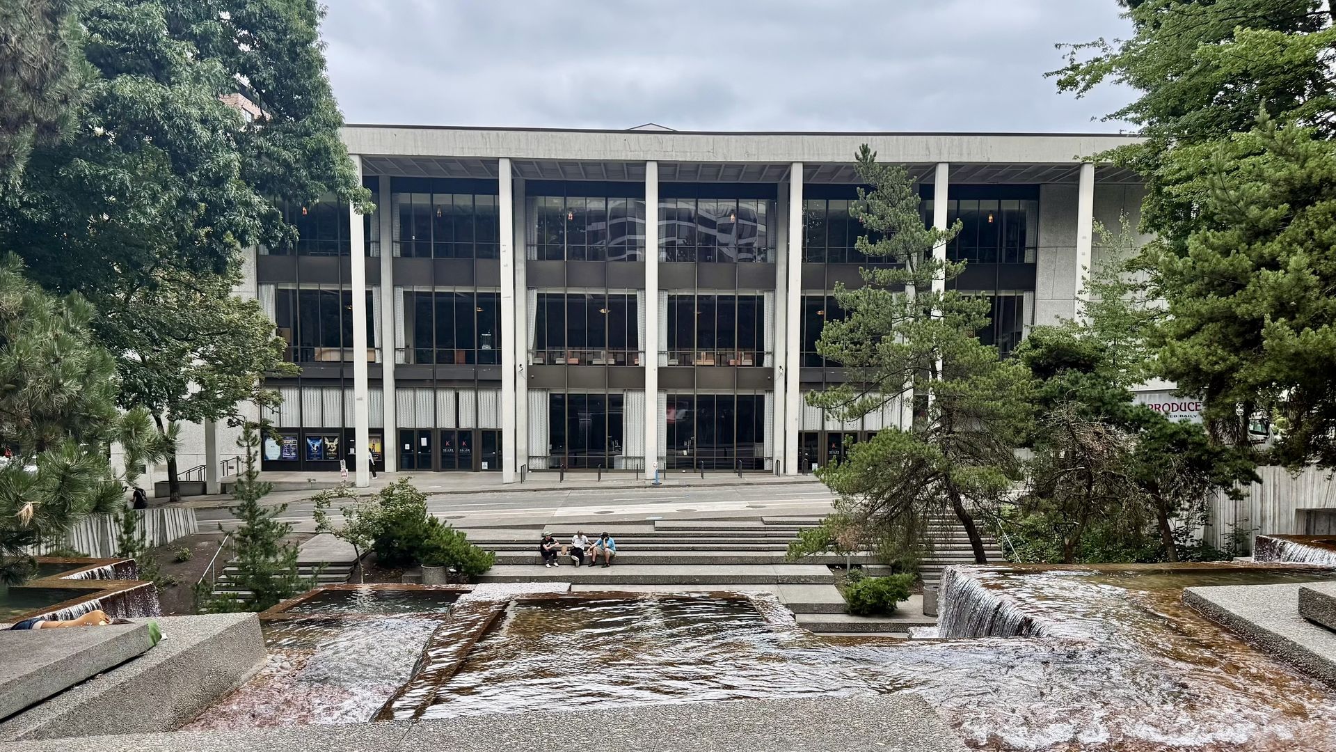 Modern glass-fronted building with tall columns, framed by green trees; a stepped plaza features a cascading water display and a few people sitting on the steps.