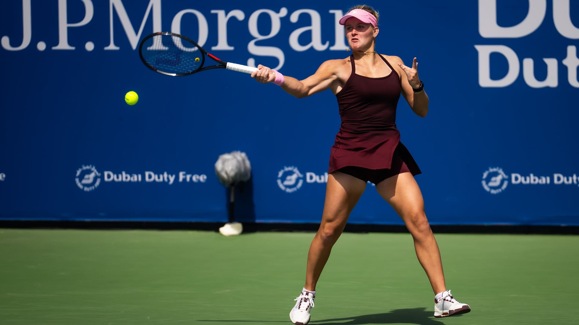Female tennis player in maroon outfit and pink visor hitting a forehand on an outdoor court with blue backdrop showing J.P. Morgan and Dubai Duty Free logos.