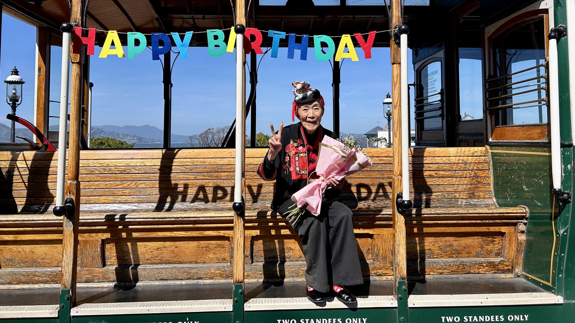 An elderly woman in a black outfit with a decorative hat sits on a wooden trolley, holding a pink bouquet and flashing a peace sign. A colorful HAPPY BIRTHDAY banner hangs above against blue sky.