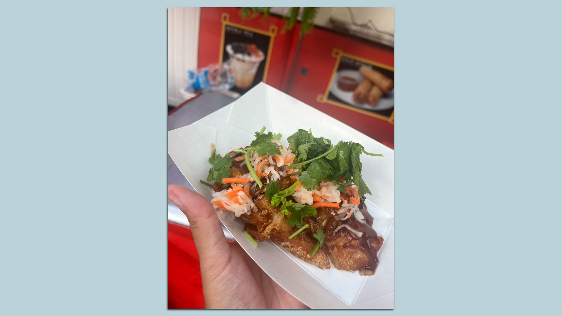 Hand holding a white tray with fried food topped with shredded carrots, white radish, cilantro, and brown sauce in front of a red background with blurry posters of drinks and spring rolls.