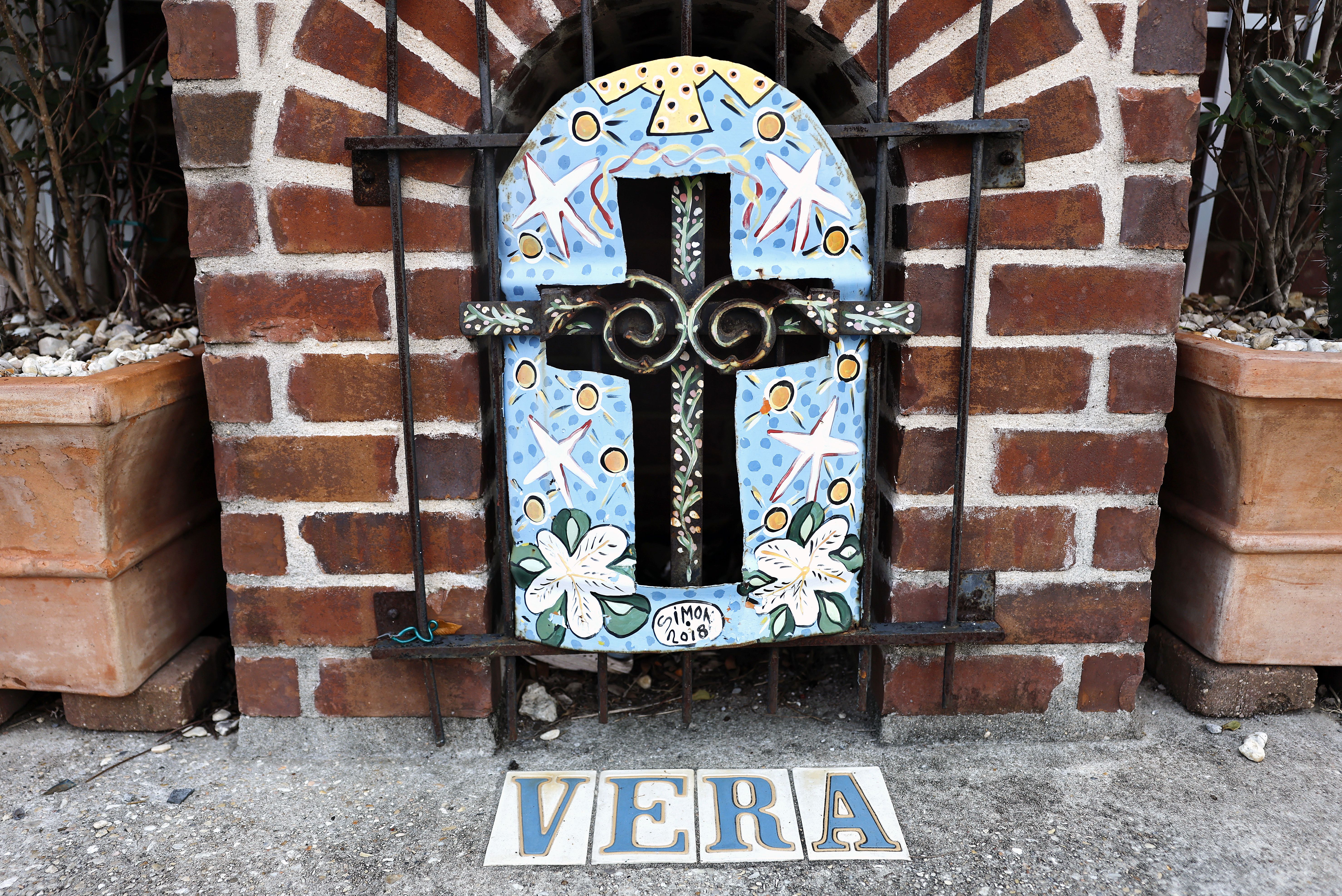 Decorative blue cross with white flowers and stars on a brick wall, secured with black metal bars, with the name "VERA" spelled out in colorful tiles on the ground below.