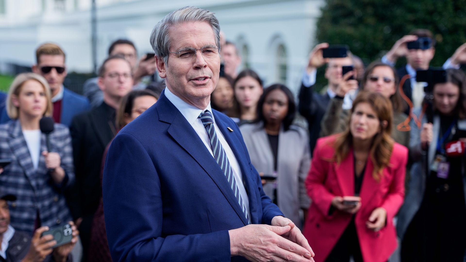 Treasury Secretary Scott Bessent, in a suit and tie and wearing glasses, addresses a group of reporters surrounding him.
