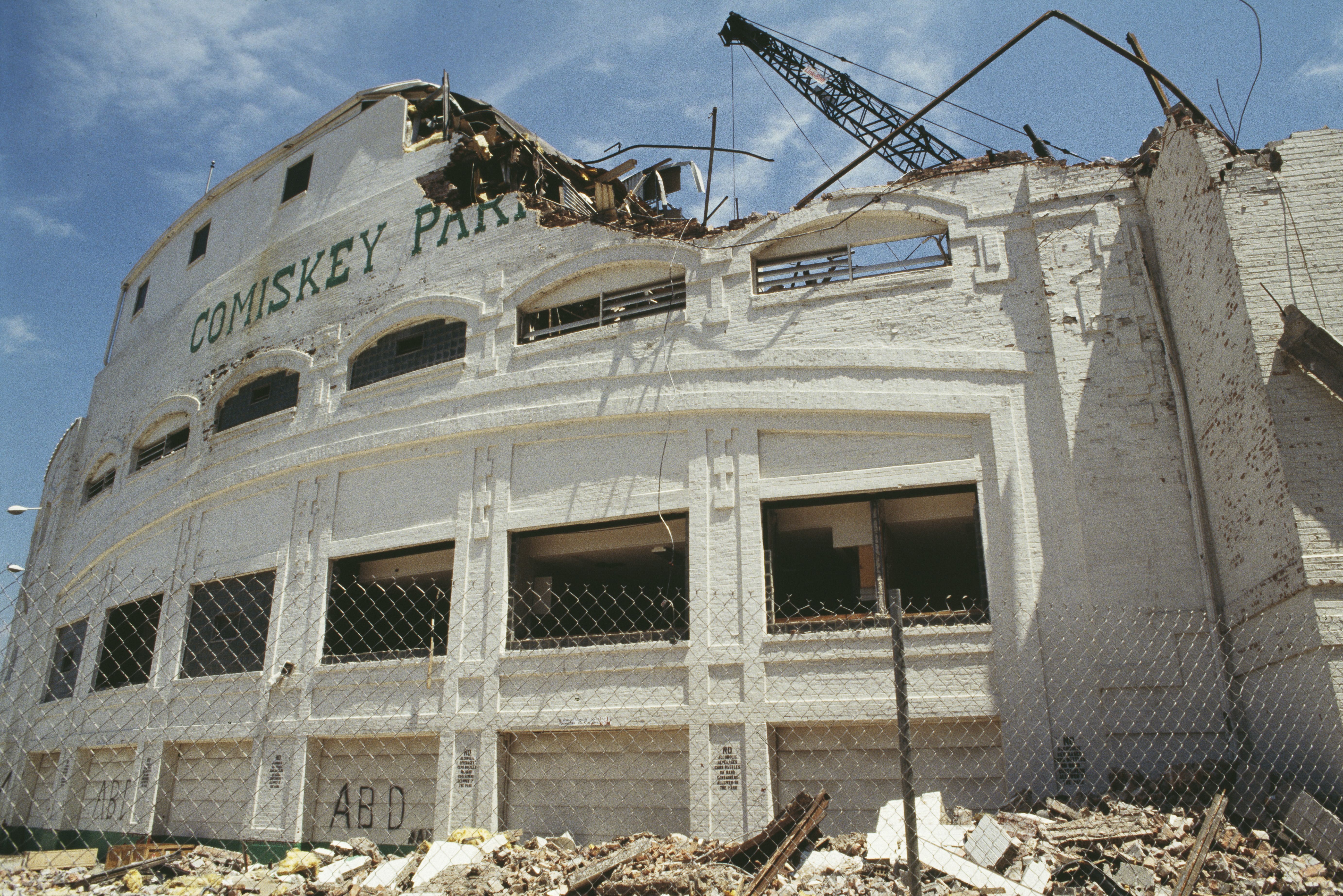 Damaged white brick facade of Comiskey Park stadium with parts of the upper wall crumbling and debris in front, under a partly cloudy blue sky and a crane overhead.