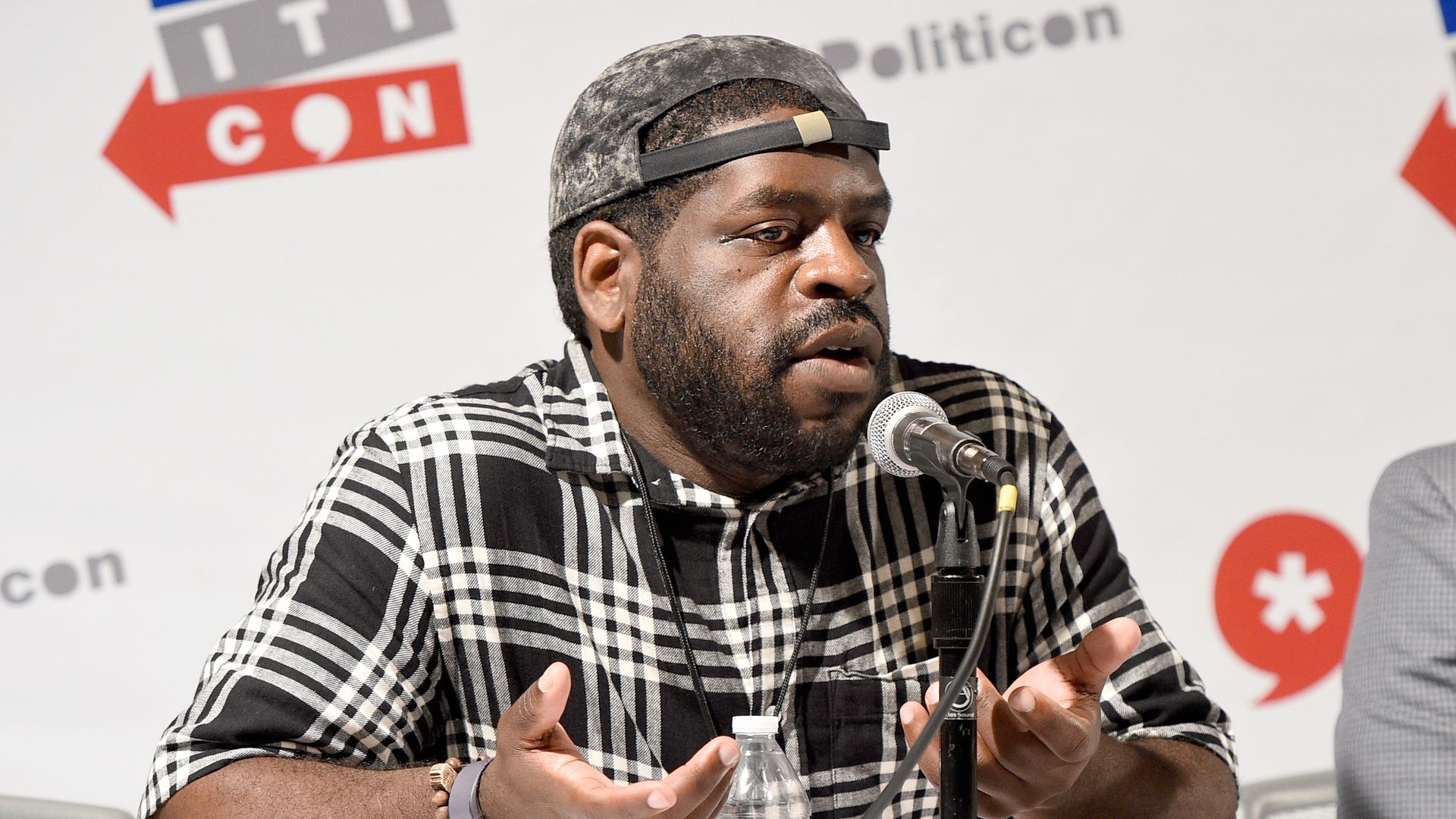 Hanif Abdurraqib speaks at a panel wearing a black and white flannel shirt and a hat.