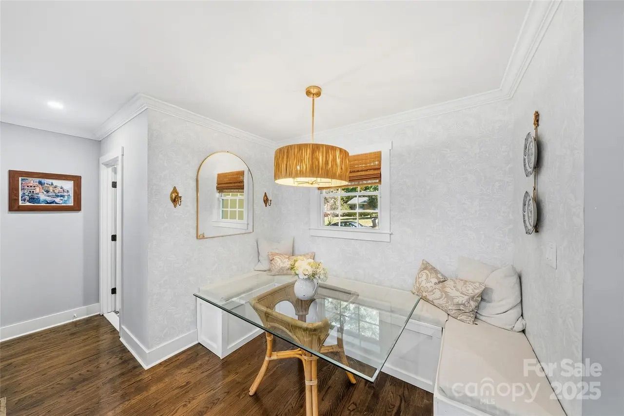 Cozy dining nook with glass table, wicker chair, built-in cushioned bench with pillows, light gray patterned walls, gold-framed mirror, wooden blinds, and hanging gold pendant light.