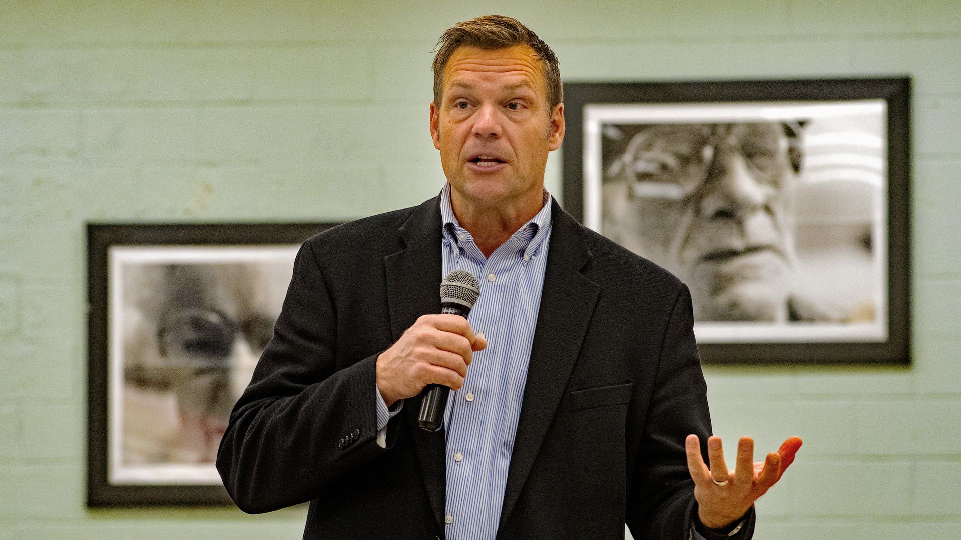 Kobach holds a microphone and wears a suit 