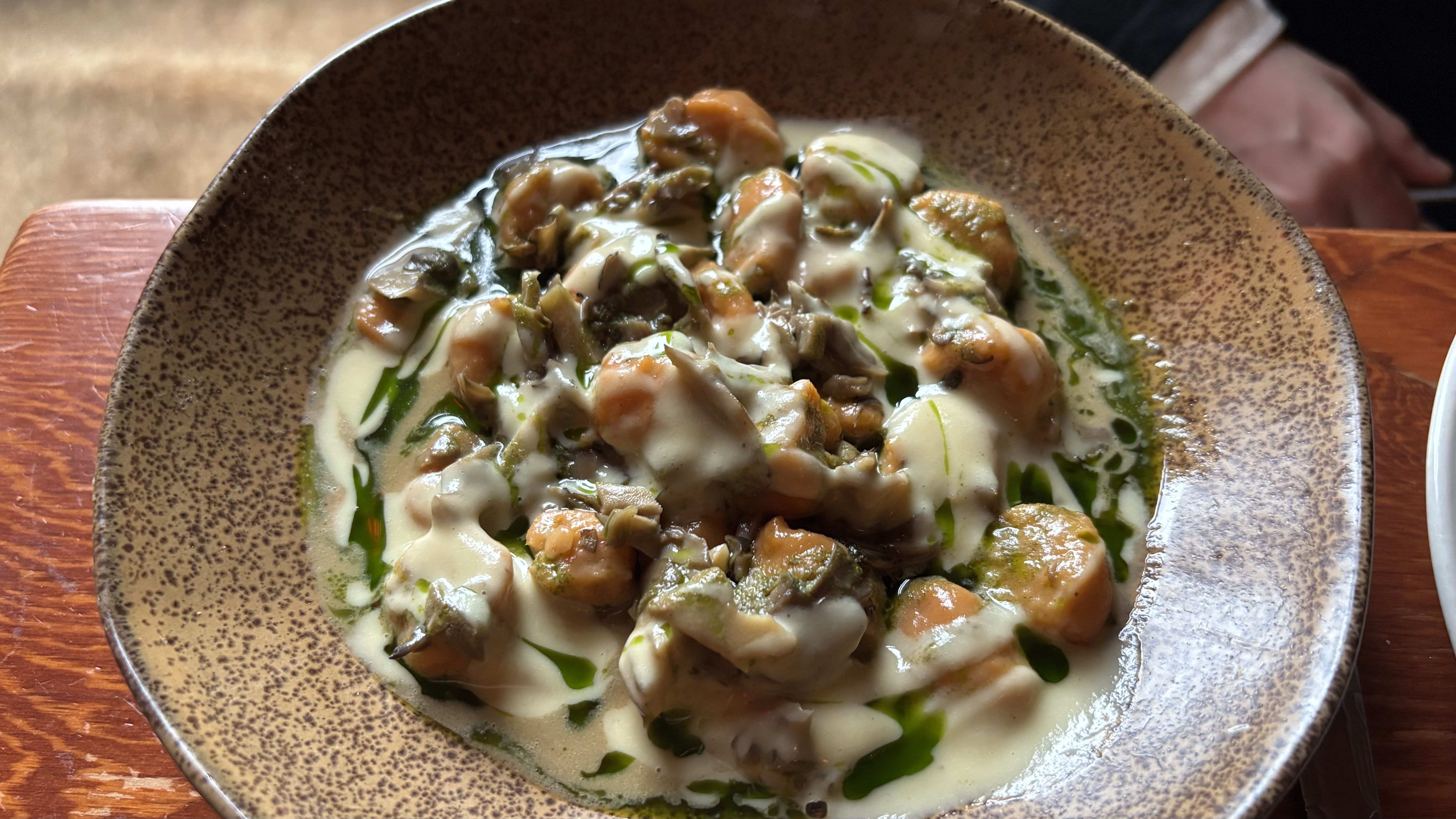 Creamy sauce with gnocchi, mushrooms, and greens in a speckled brown bowl on a wooden table; a hand is visible in the background.