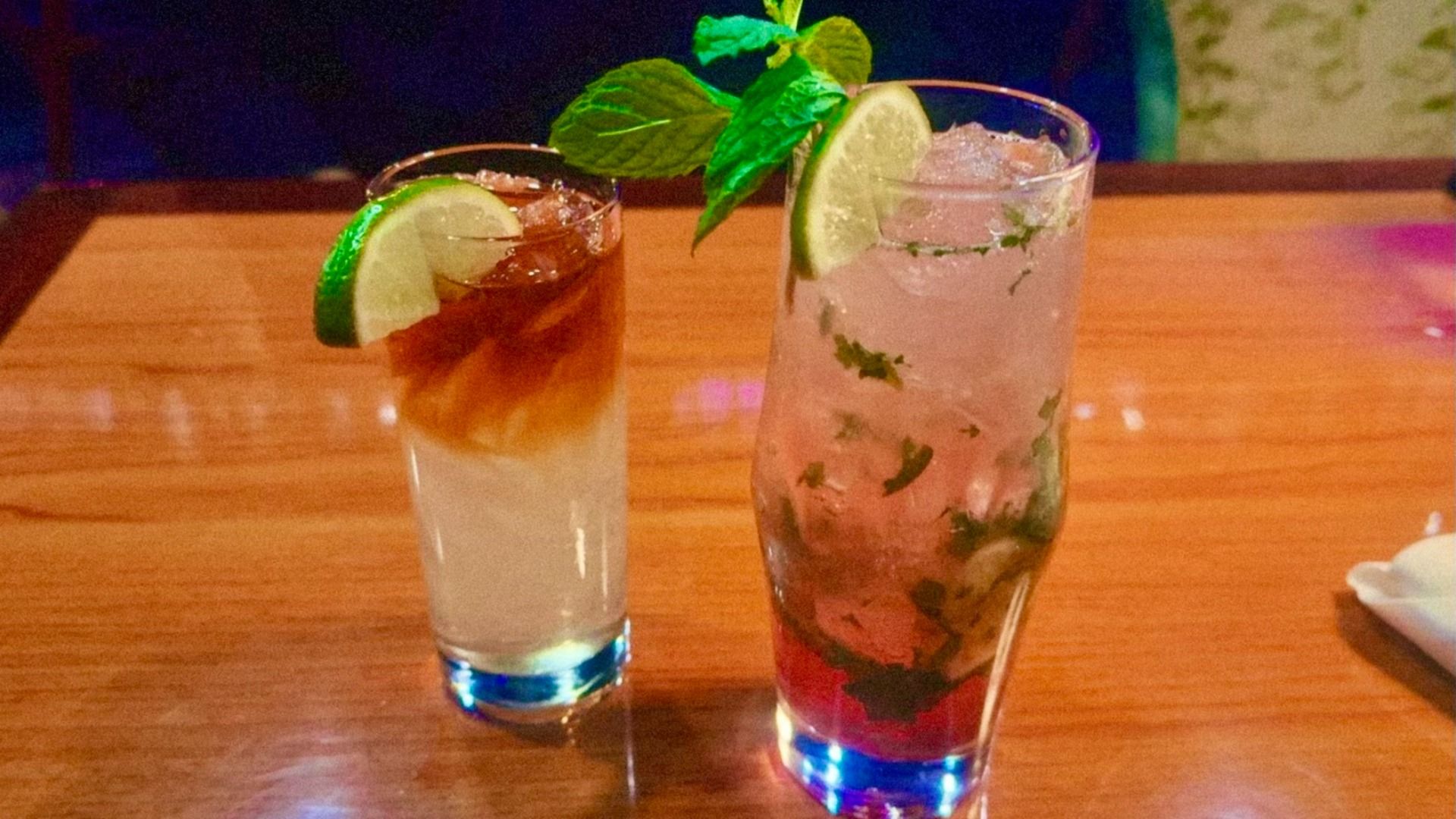 Two iced cocktails on a wooden table, one with lime and dark top layer, the other with lime, mint leaves, and a reddish bottom layer.