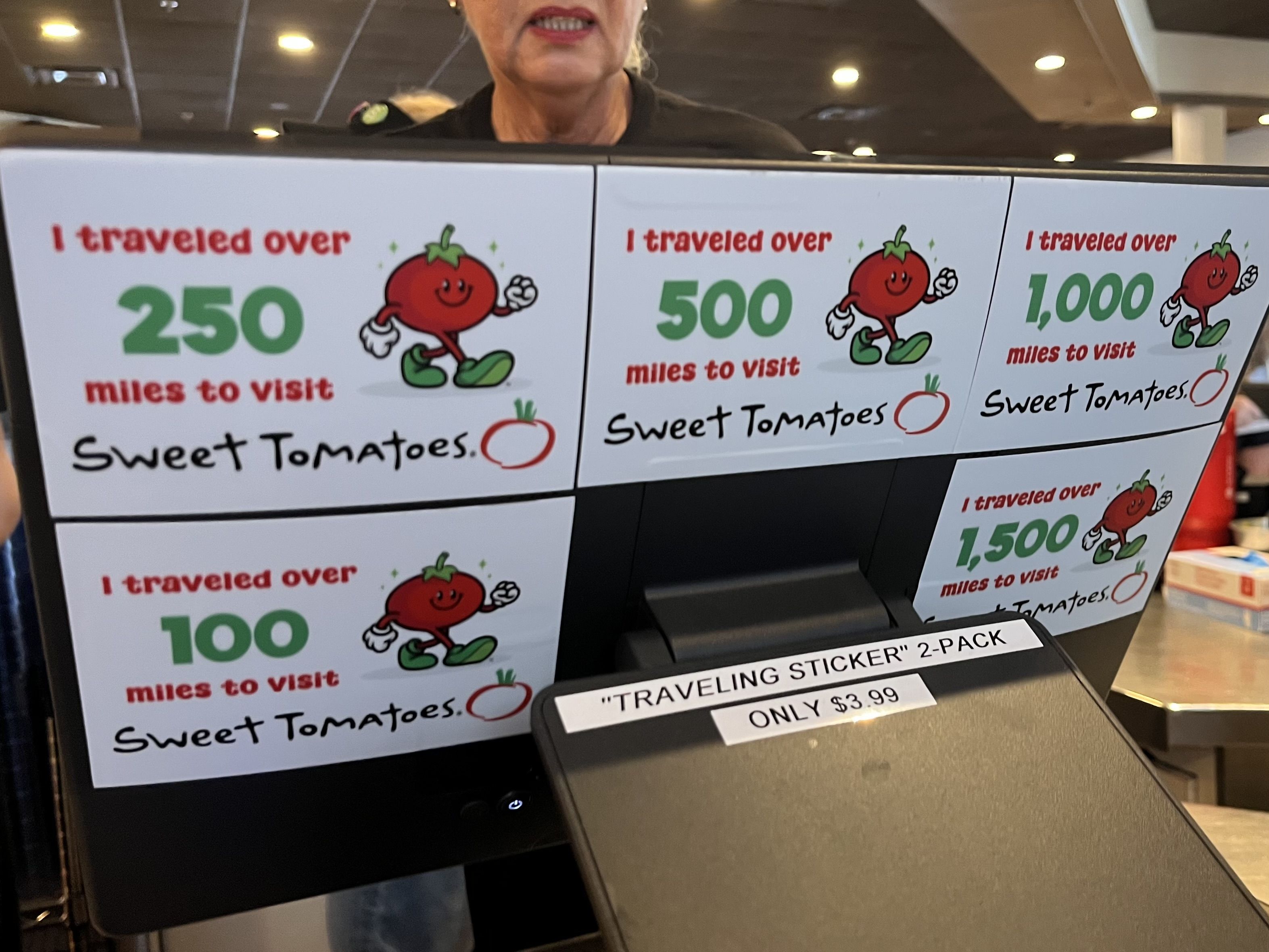 Display wall with posters showing a smiling red tomato mascot, the phrase "I traveled over" and large green mile numbers (250, 500, 1,000) to visit, plus "Sweet Tomatoes" at the bottom.