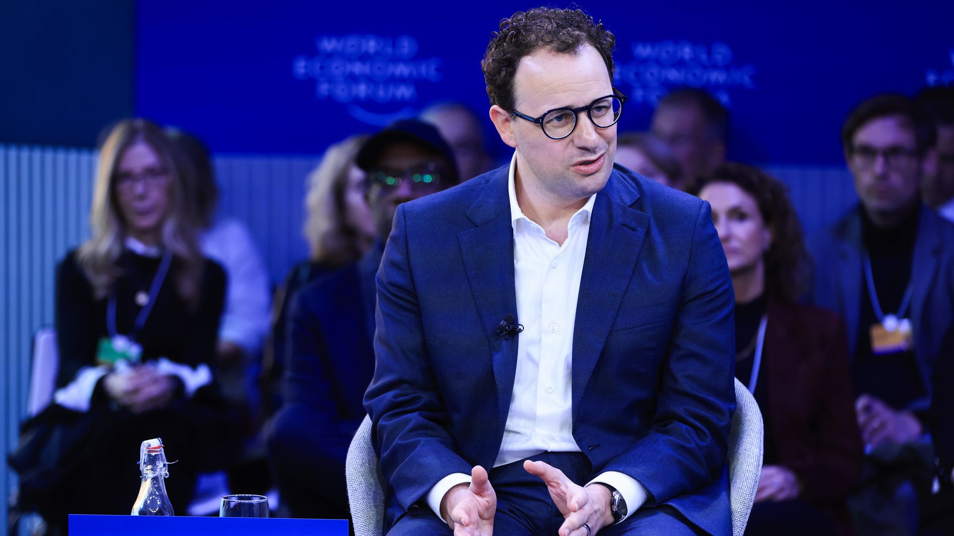 Person in a dark blue suit and glasses speaking at the World Economic Forum, seated with hands gesturing, with a nameplate reading "AMODEI" in front and a blurred audience behind him.
