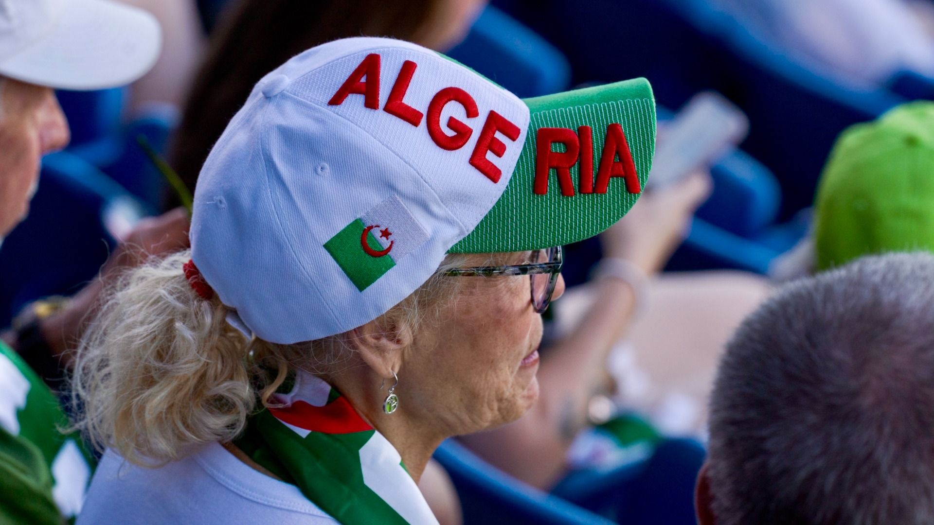 Close-up of a spectator wearing a white cap with red ALGERIA lettering and a green Algeria flag patch; the person has glasses, blonde hair, and a green-striped scarf at a crowded stadium.