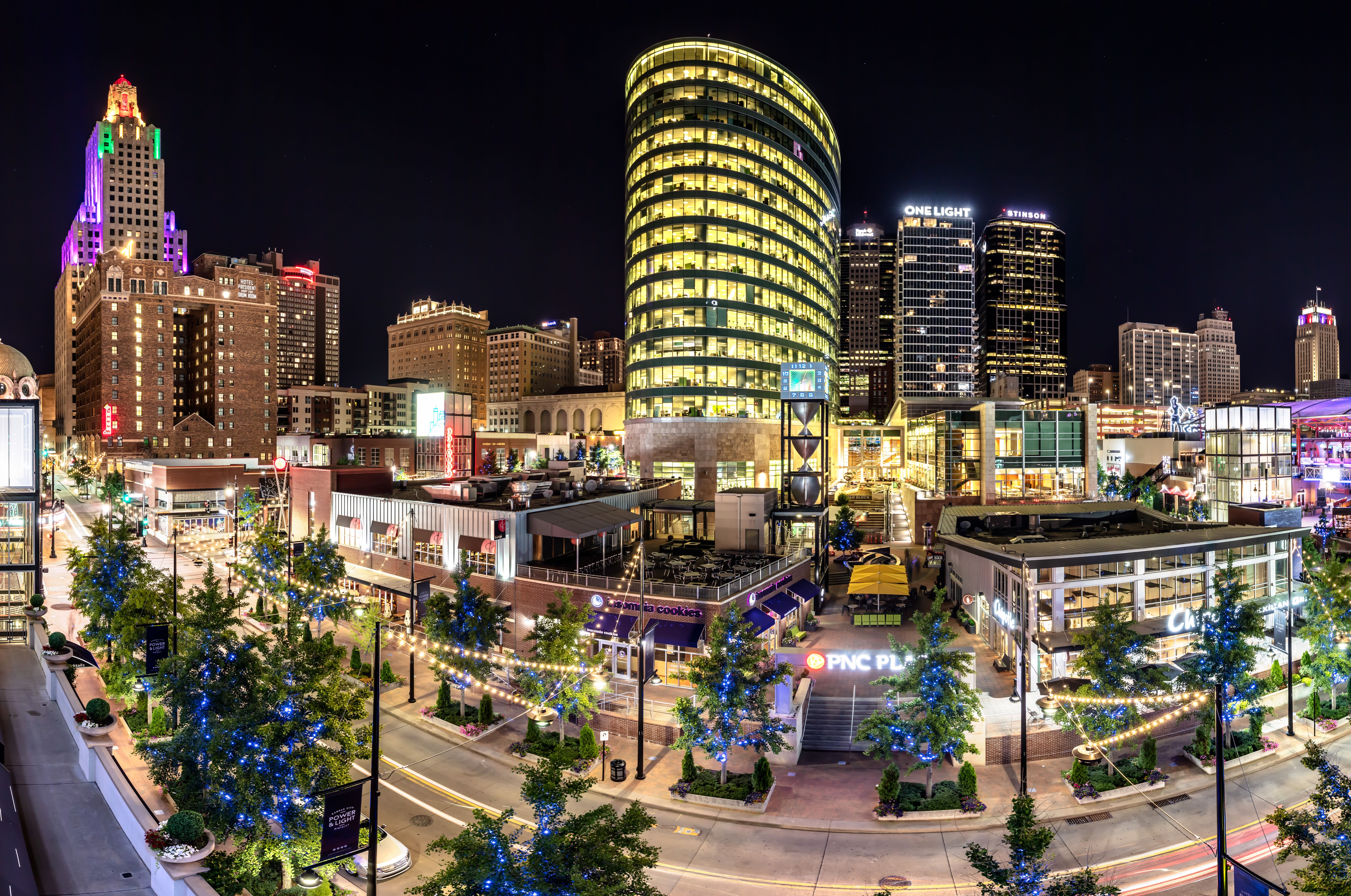 Nighttime cityscape with brightly lit modern and historic buildings, trees wrapped in blue and white string lights, and streets adorned with warm yellow hanging lights.
