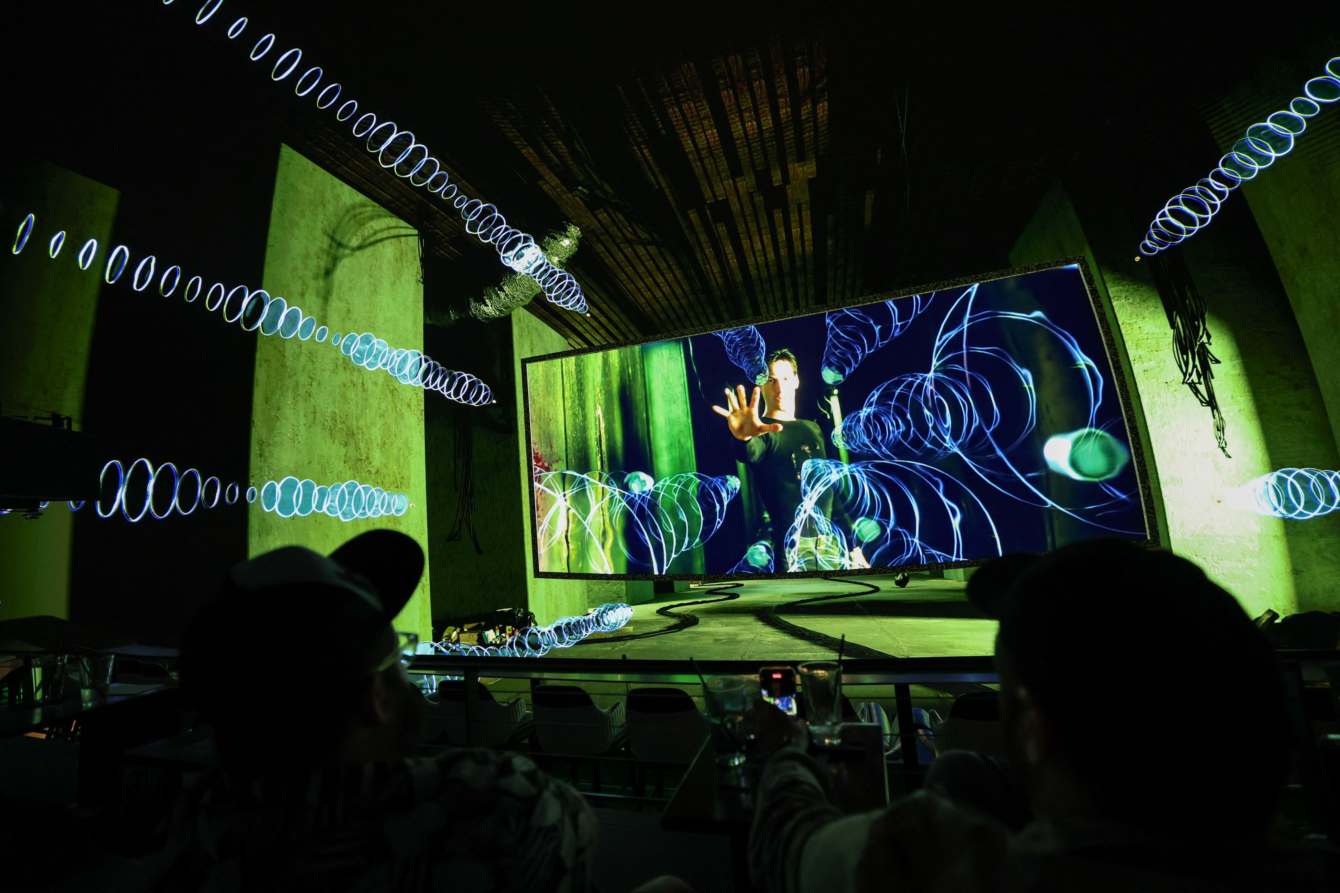 People watch a scene from a movie projected on a large screen in a dark room with green walls and blue spiraling light effects in the foreground.