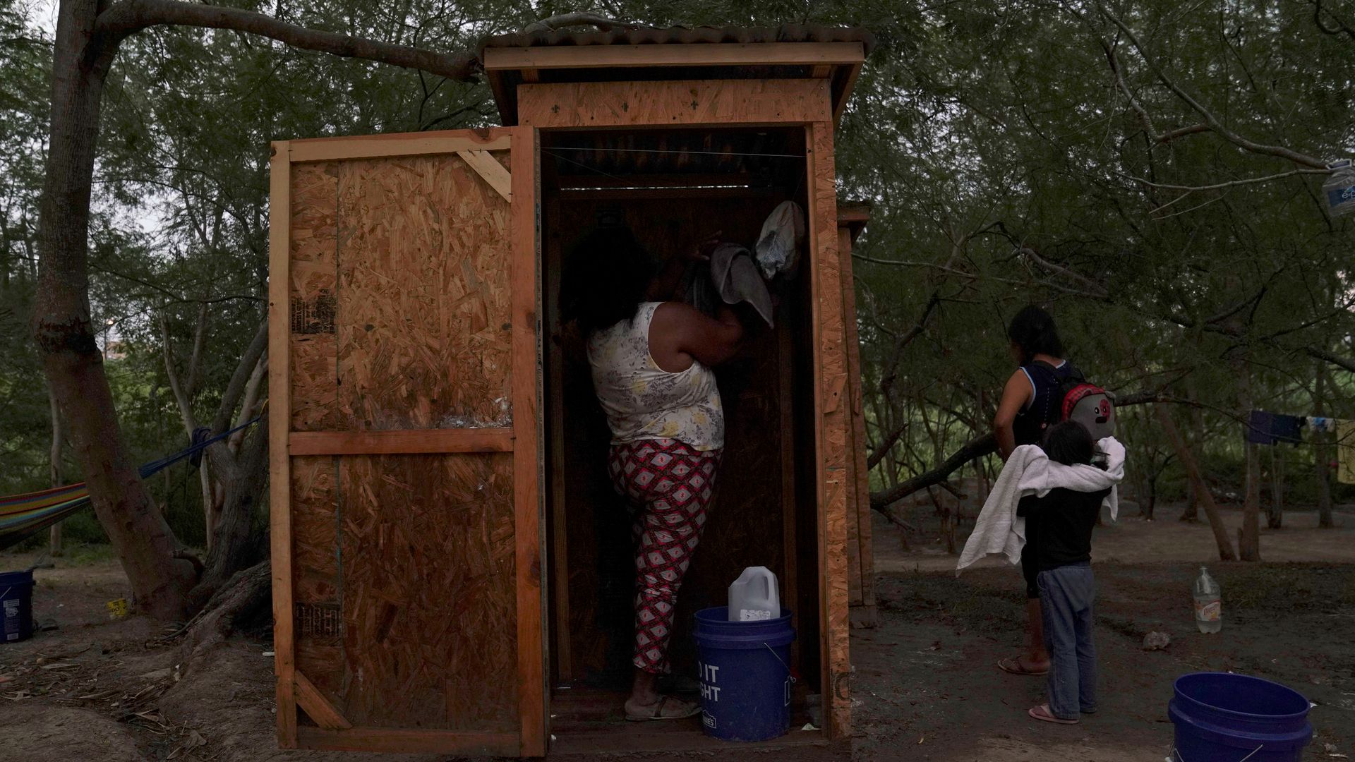 This image shows a woman standing in leggings and a tank top in a wooden outdoor shower cubicle. There are buckets outside the cubicle.