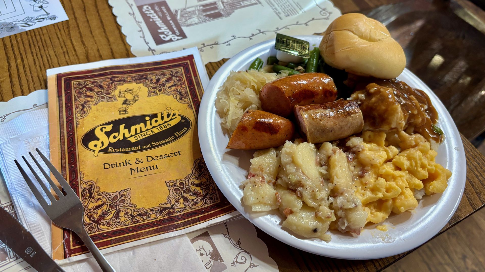 A heaping plate of buffet food — including sausages, a roll, German potato salad and macaroni and cheese — next to a Schmdit's menu and a fork.