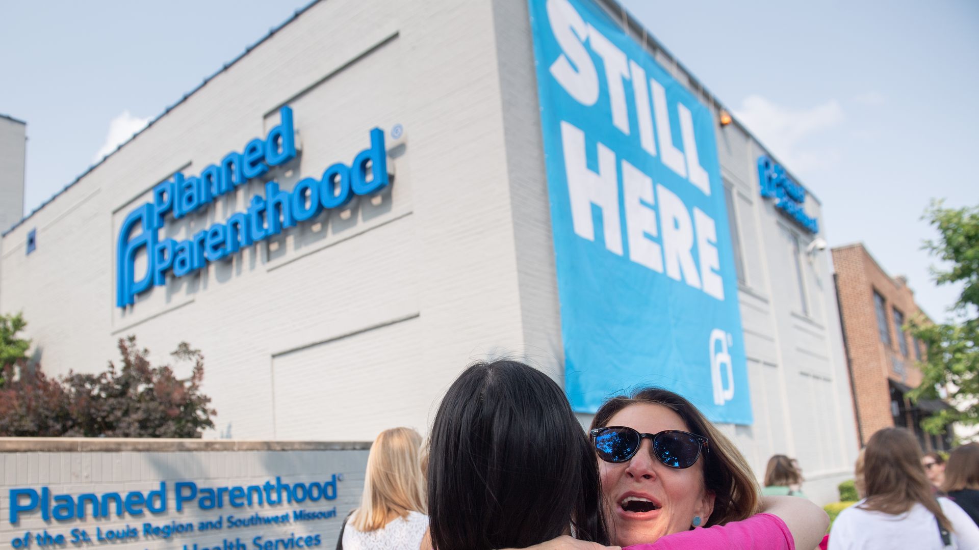 In this image, two women in bright shirts hug outside of a Planned Parenthood with a large banner that reads "still here."