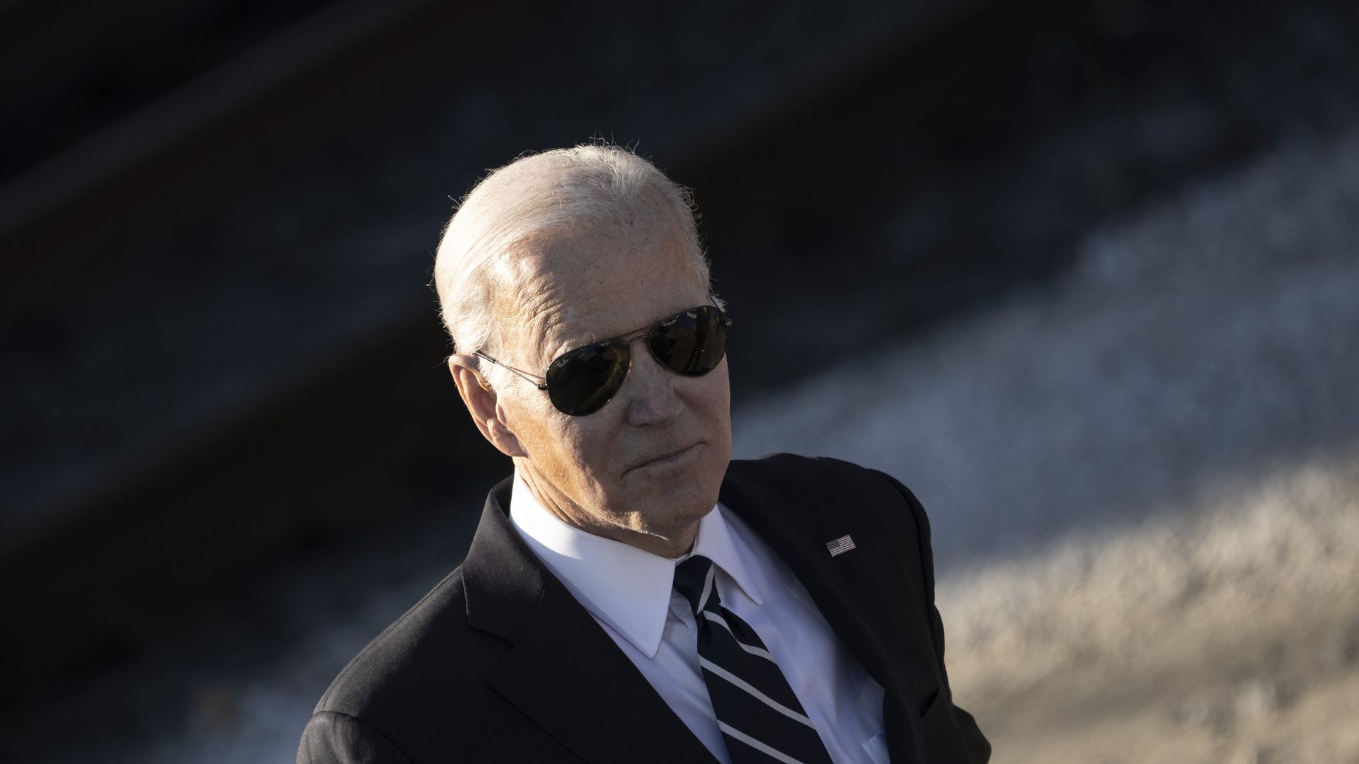 President Biden, wearing a gray suit, aviator sunglasses, white shirt and white and navy striped tie stands in front of train tracks.
