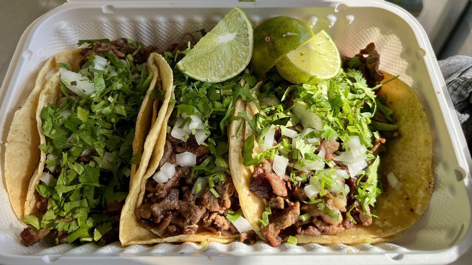 Three tacos in a white takeout container topped with grilled meat, chopped onions, cilantro, and two lime wedges.