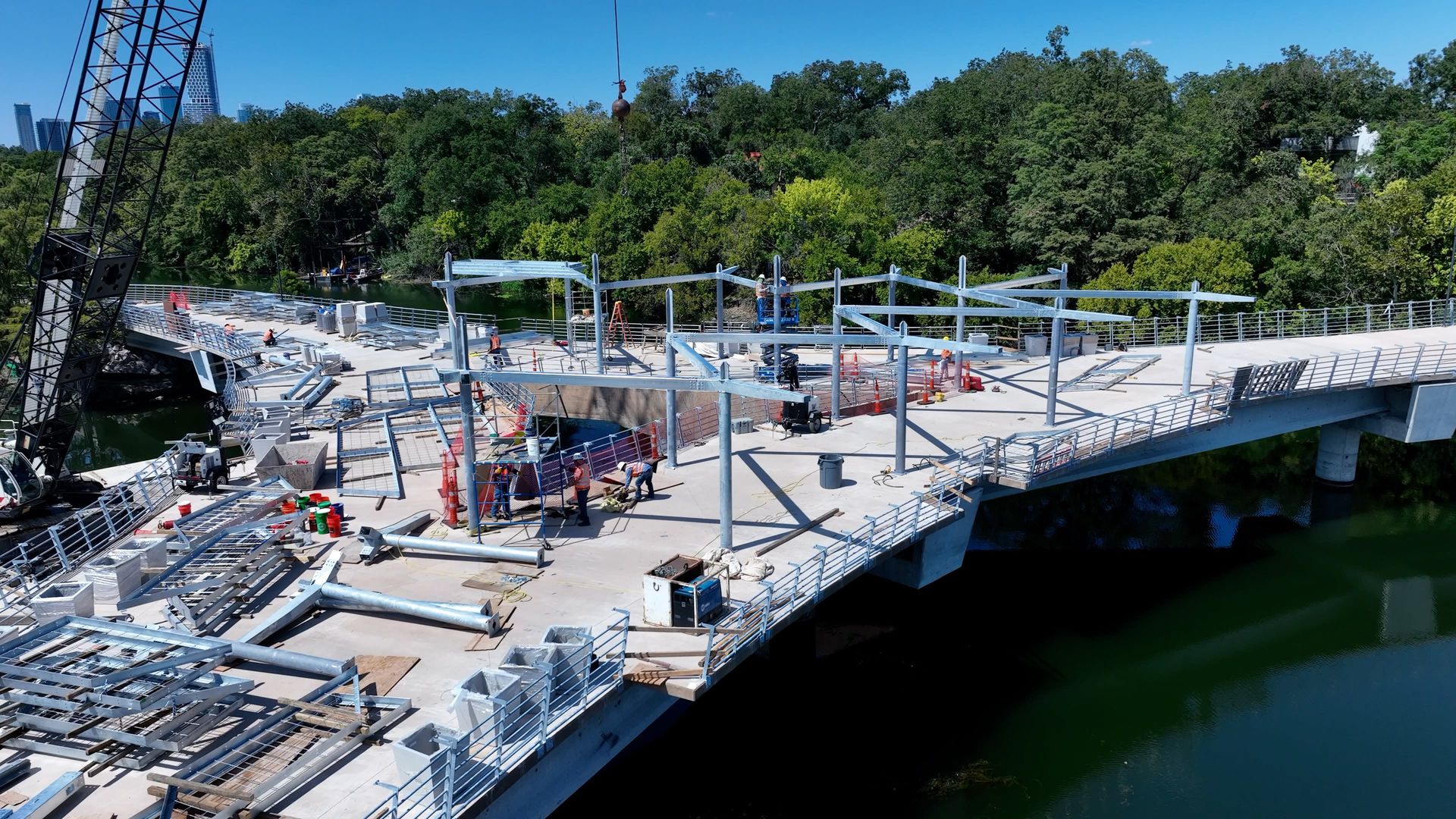 Construction workers assembling metal framework on a wide bridge over a river, surrounded by green trees and blue sky in the background.