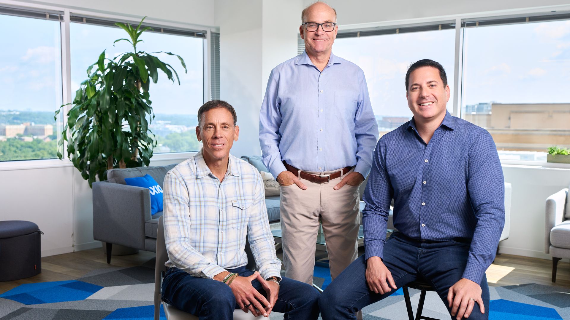 Three men in an office pose for a photo: center man in a light blue shirt stands, flanked by two men seated in plaid and blue shirts. Large plant, gray furniture, and windows with a city view.