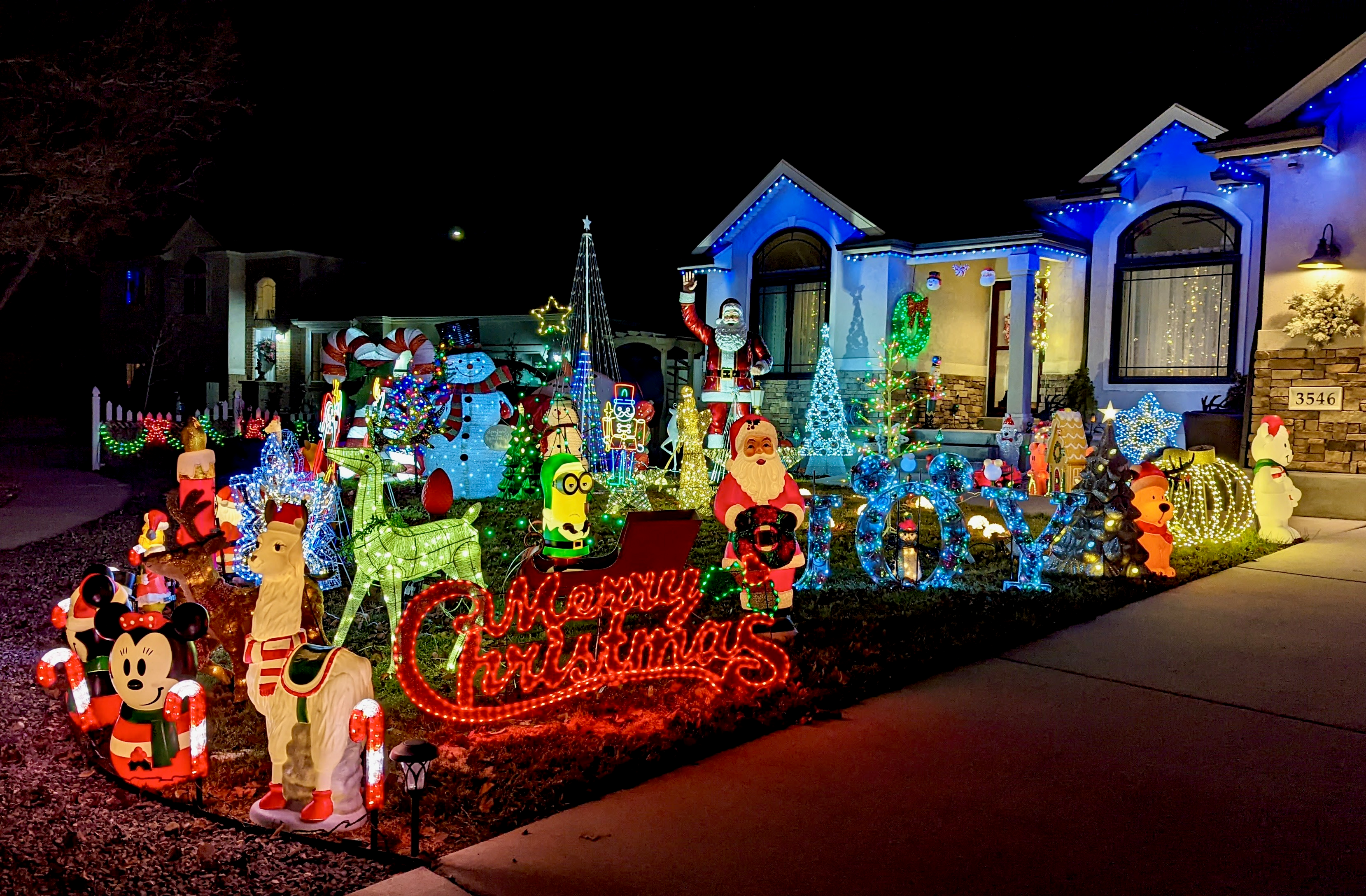 A christmas lights display with lots of figurines and a red "Merry Christmas" sign in a residential yard 
