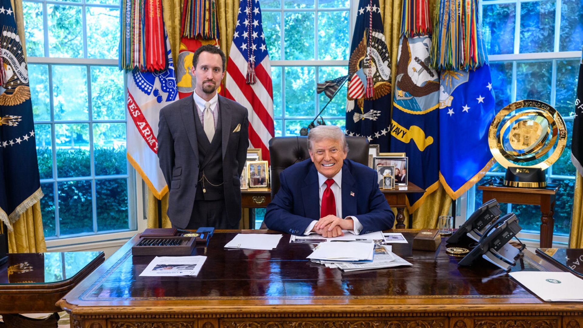 Two men in the Oval Office; one, President Trump, seated behind the desk wearing a navy suit and red tie, smiling, the other standing in a dark suit and tie. US flags and military banners in background.