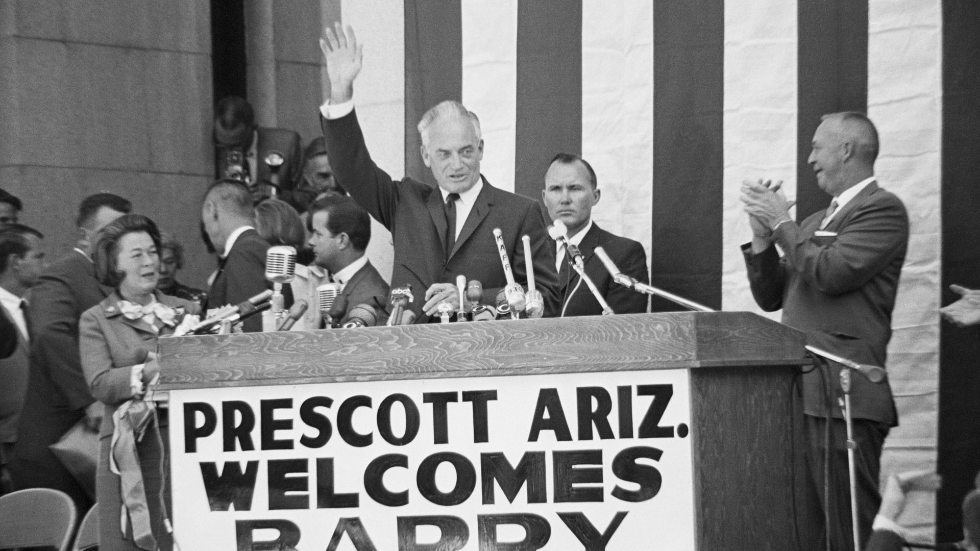 Black and white photo of Barry Goldwater waving while standing in front of a large lectern filled with microphones.