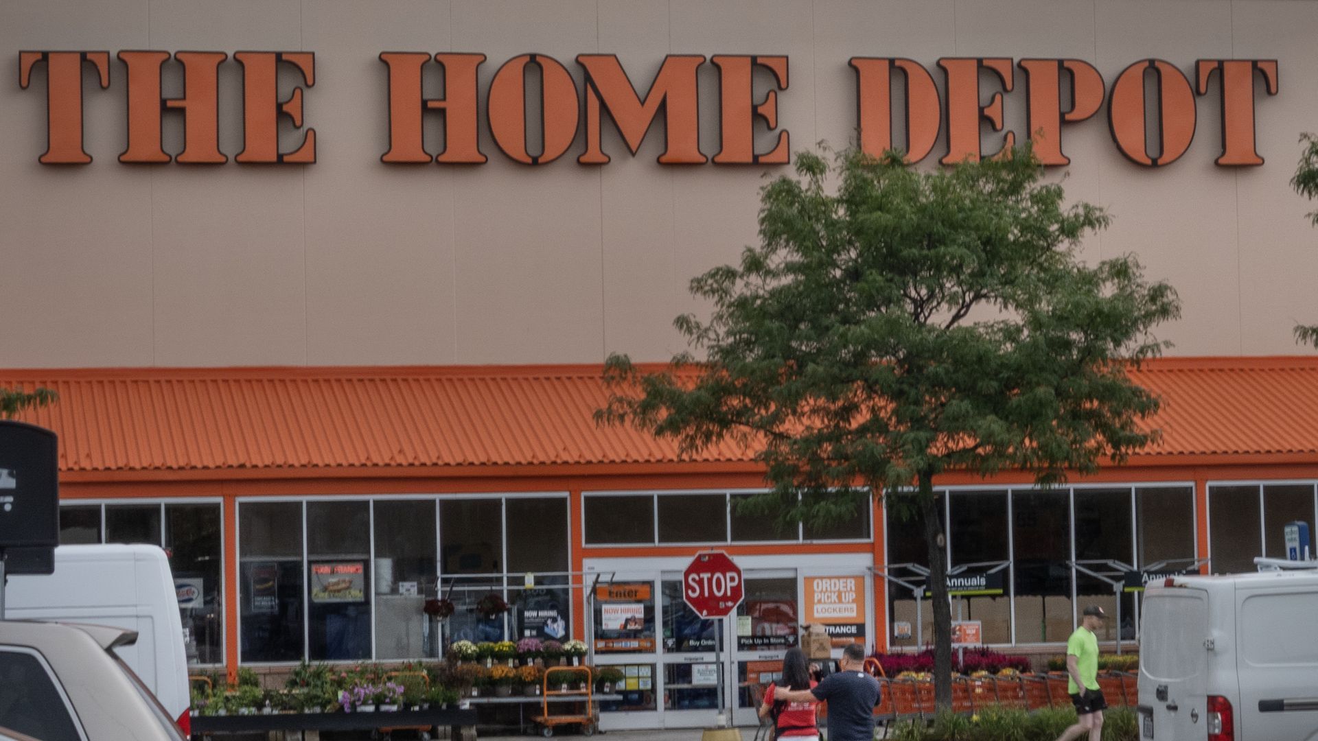 Exterior of The Home Depot store with orange signage, orange awning, a tree, people walking, and a stop sign in front.