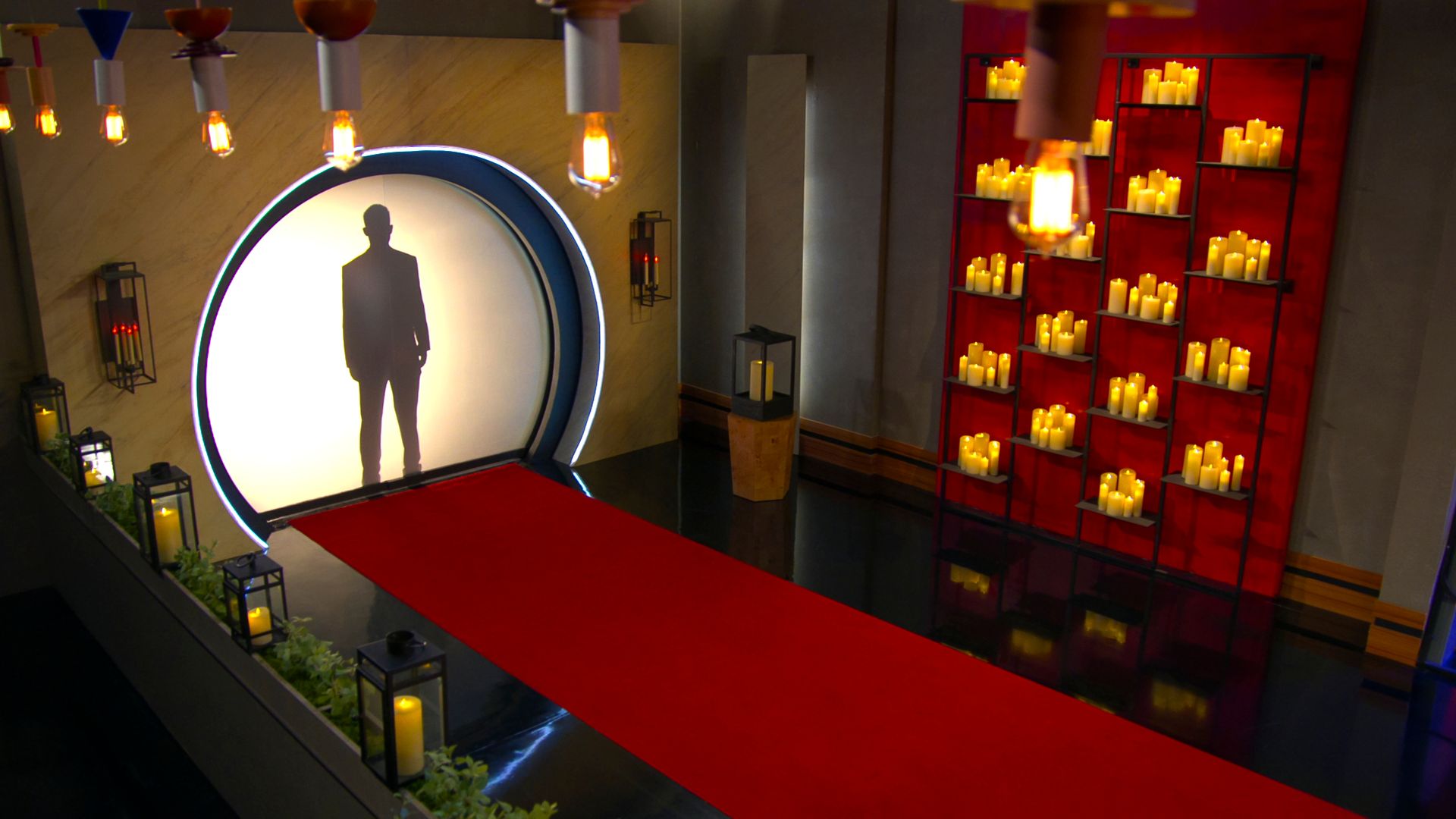 A silhouette of a person stands behind a circular white backdrop with blue lighting edges. A red carpet leads up to the backdrop, surrounded by candle lanterns and a red panel filled with lit candles.