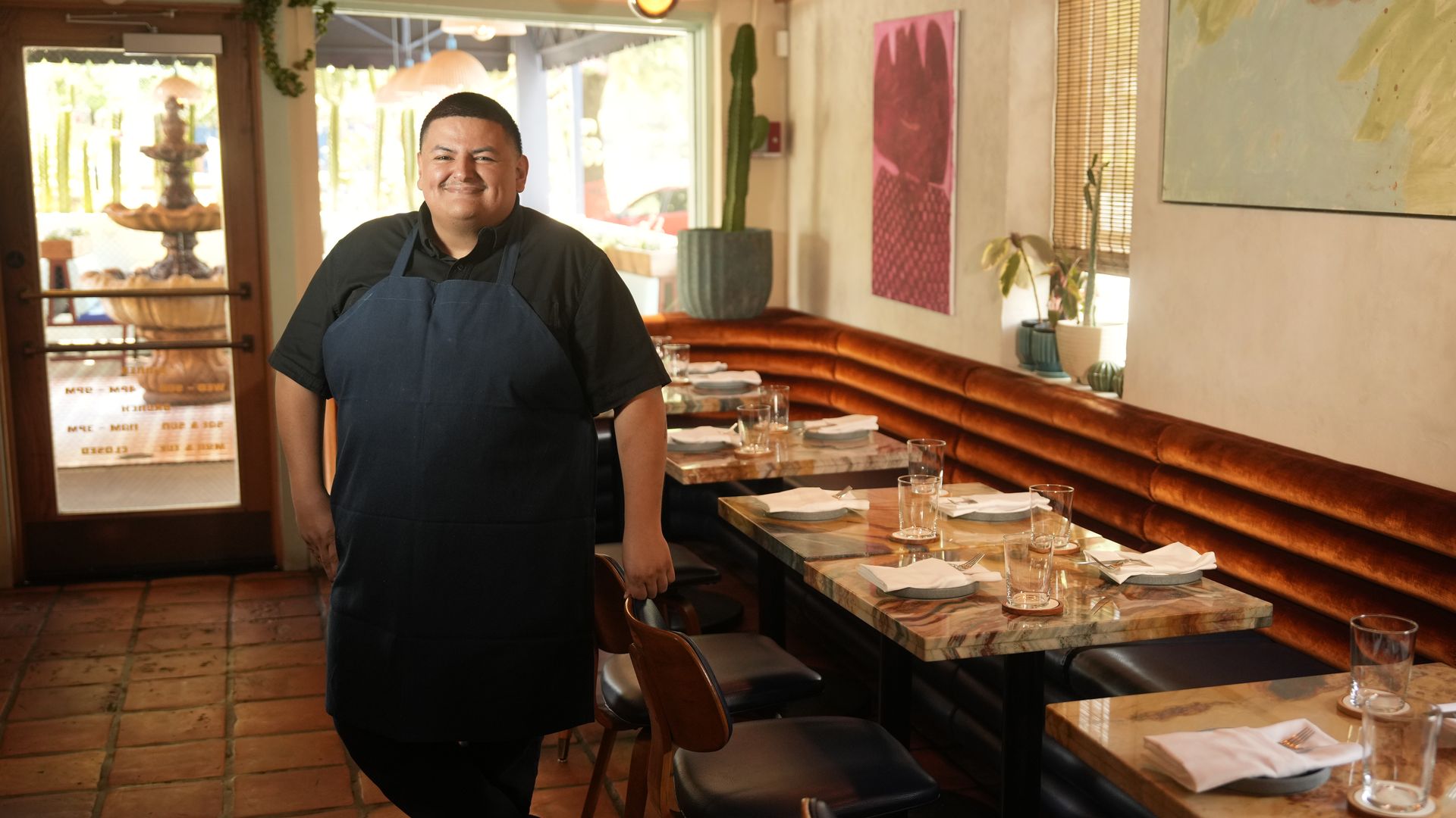 Smiling chef in a dark shirt and blue apron stands beside marble-top tables set with napkins, glasses, and cutlery inside a cozy, sunlit restaurant with orange-brown bench seating and potted plants.