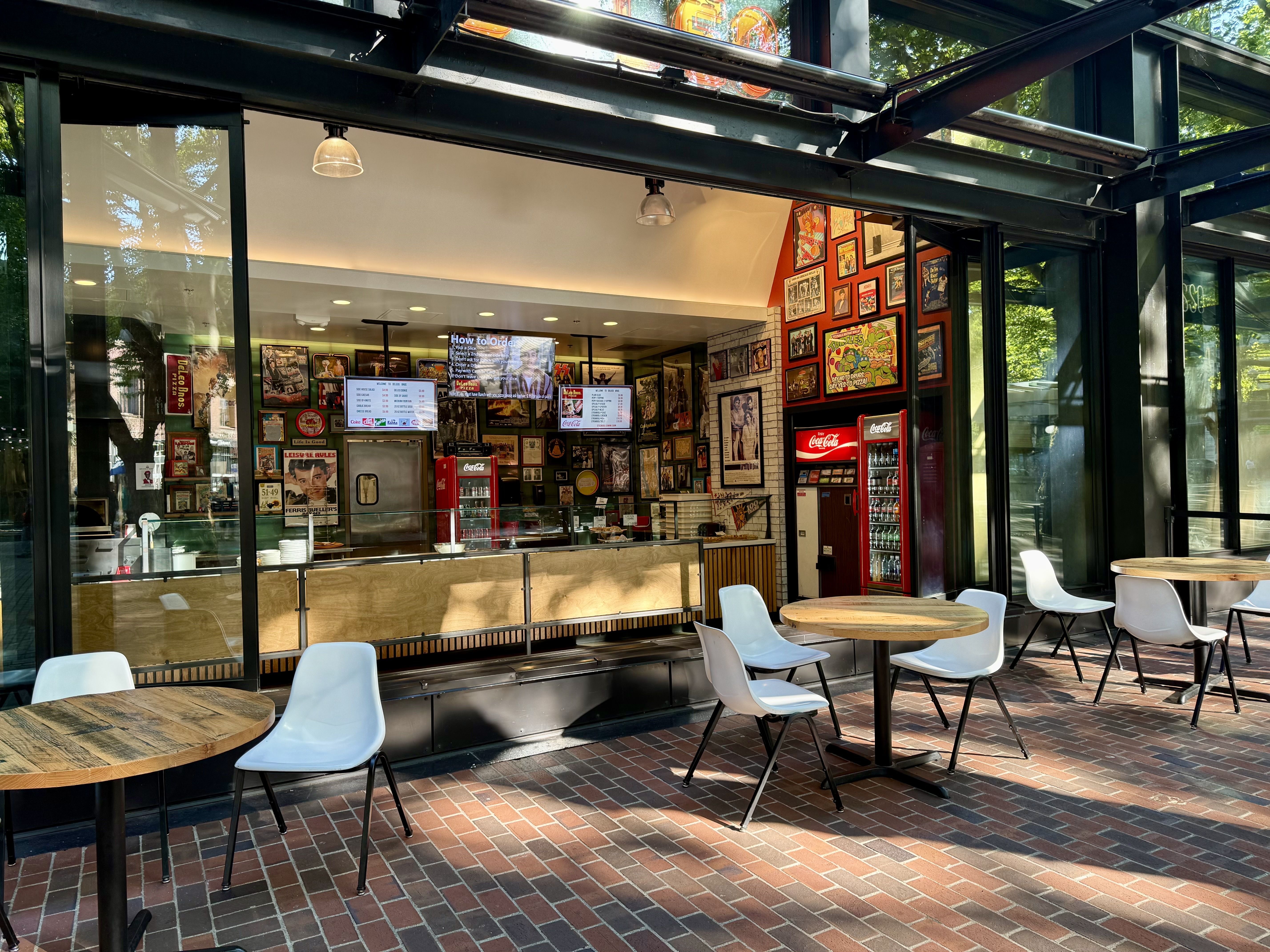 Outdoor seating area with wooden round tables and white chairs in front of a restaurant with large glass windows displaying framed posters, Coca-Cola fridge, and digital menu screens.