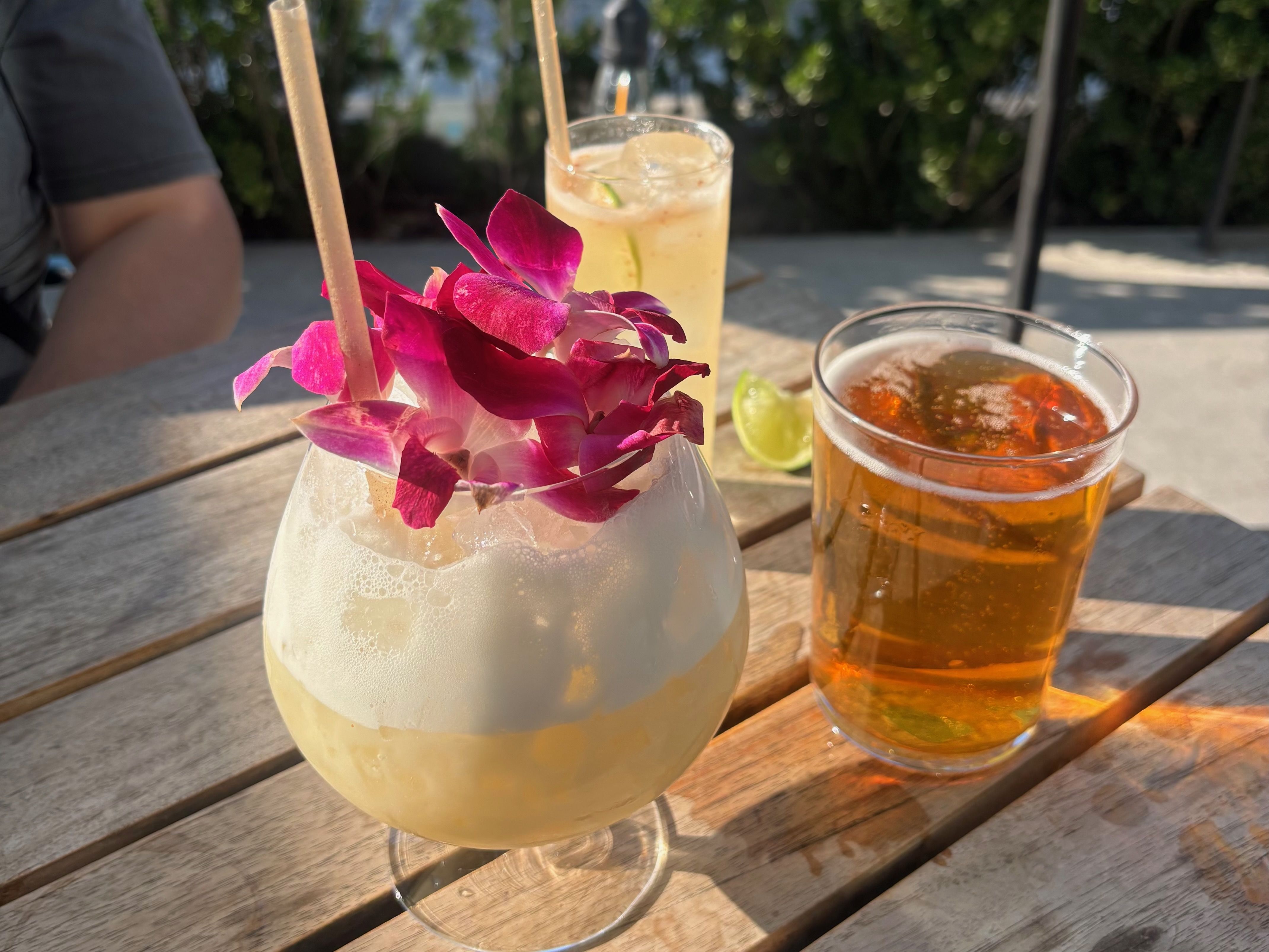 Three drinks on a wooden table: a frothy pale cocktail with pink orchid flowers, a clear glass of amber beer, and a tall iced yellow drink with lime wedges in the background outdoors.