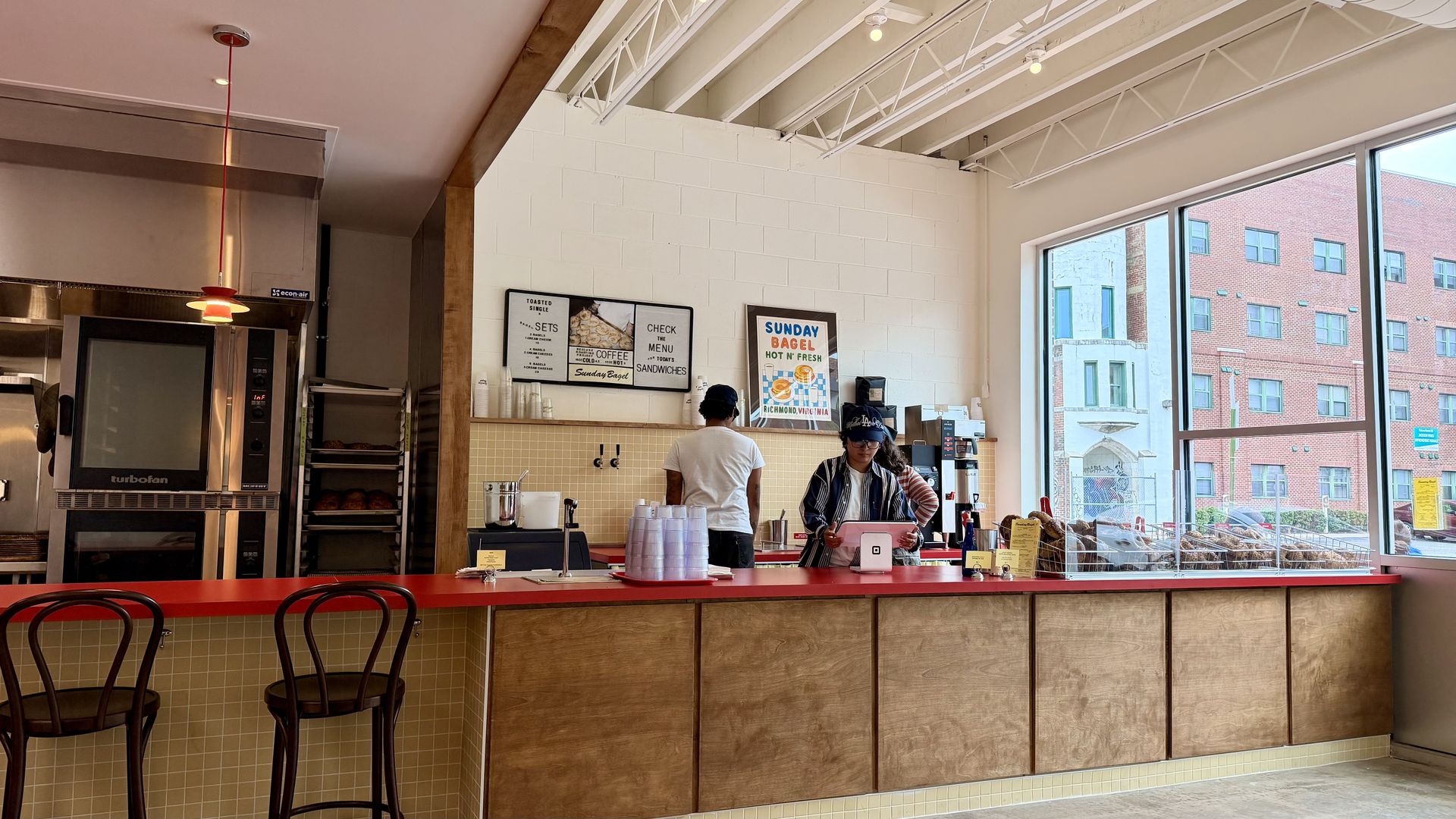 Bright cafe interior with a red counter and white brick wall. Two staff at the counter prepare drinks. Large windows show a brick building outside; pendant light and metal ceiling trusses above.