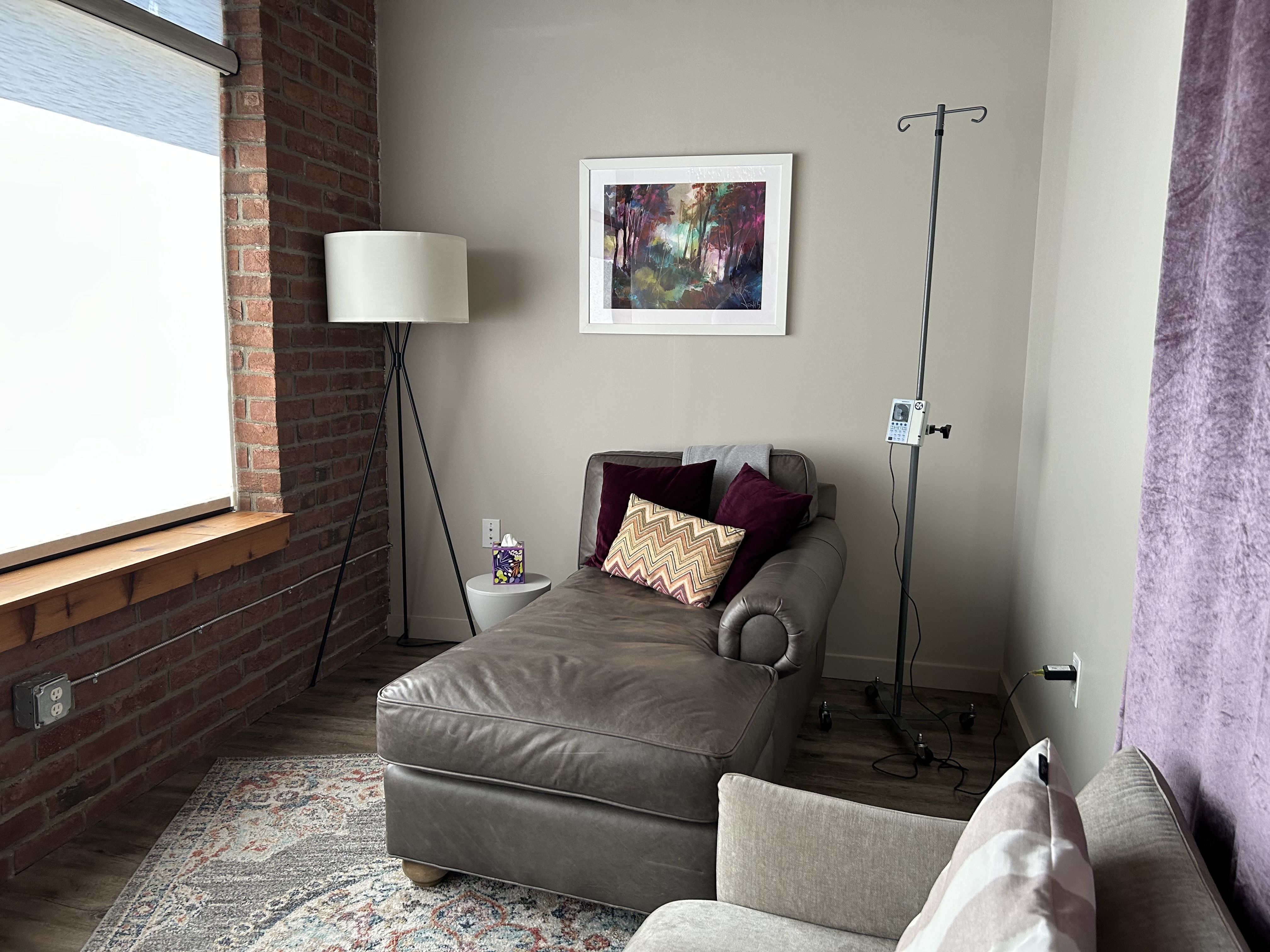 Cozy living room with an exposed brick wall, a gray sectional sofa with burgundy pillows, a white-framed abstract painting, a tall floor lamp, an IV pole with pump, purple curtains, and a rug.