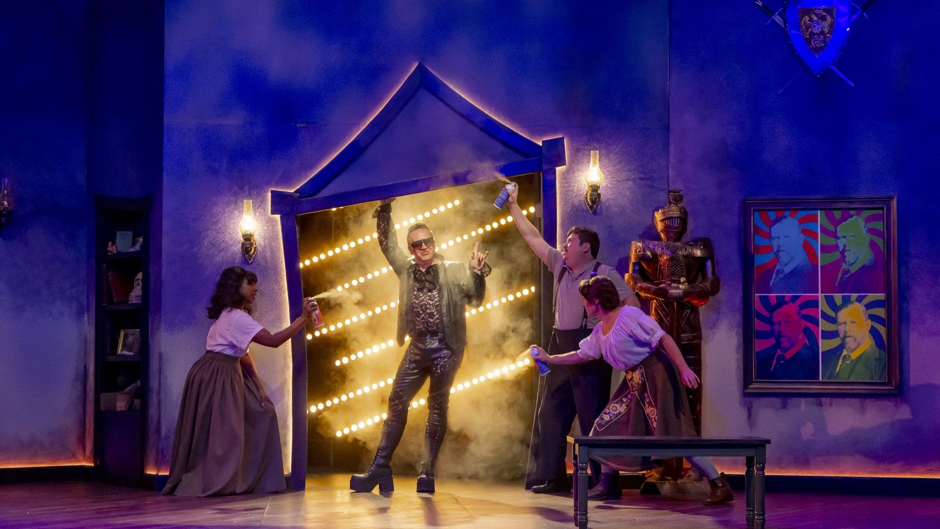 Blue-lit stage with smoke. A man in a shiny black leather outfit raises his arms inside a lit doorway; others dance and spray cans. A shield with swords and colorful wall art are visible.