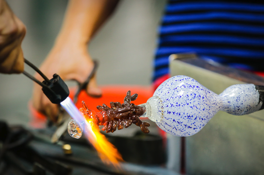 Photo shows two people working on a blown glass creation with a sea creature on it.