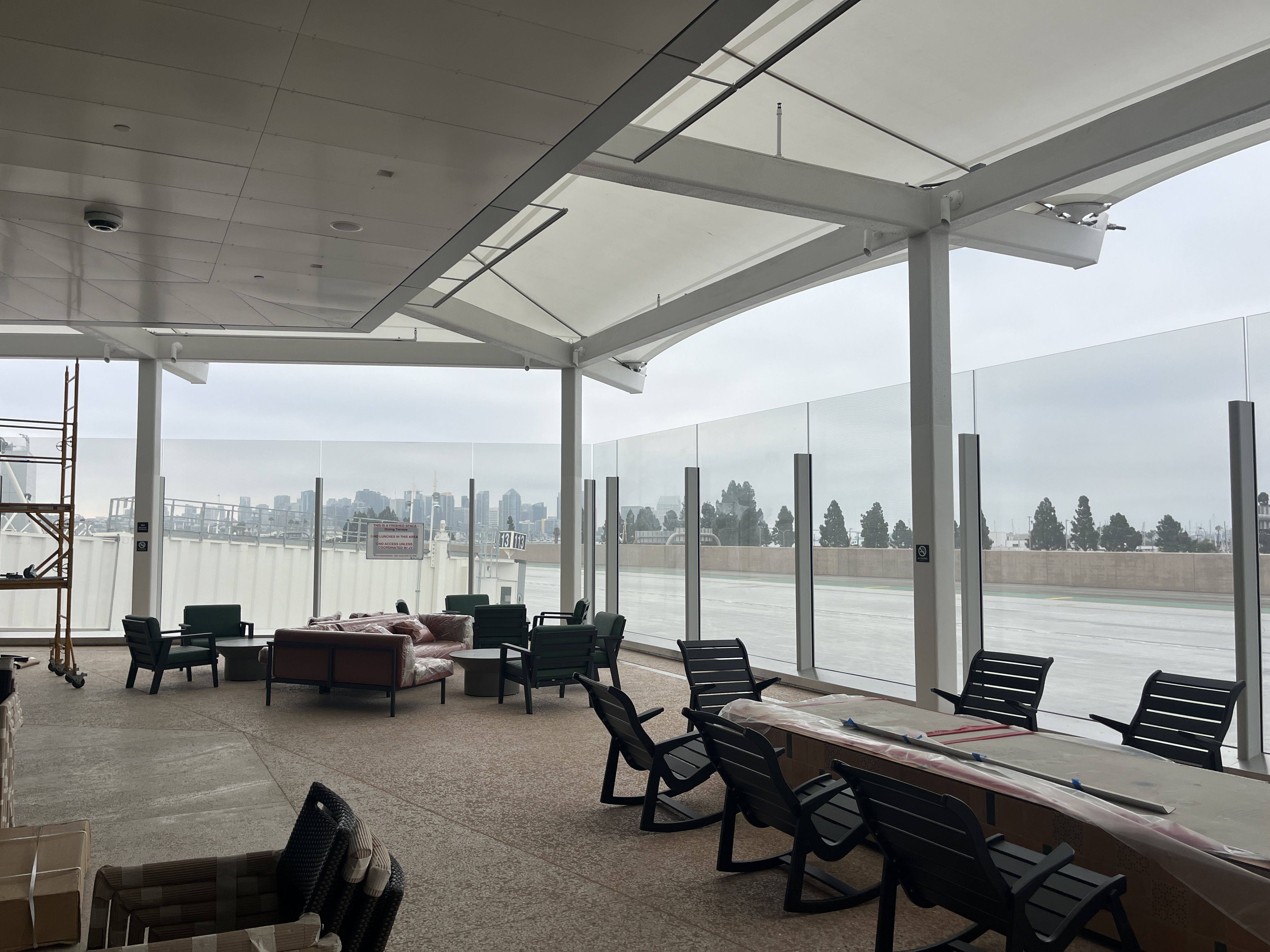 Covered outdoor patio with black chairs, couches and tables, some wrapped in plastic, overlooking a foggy San Diego skyline behind glass panels on a cloudy day.