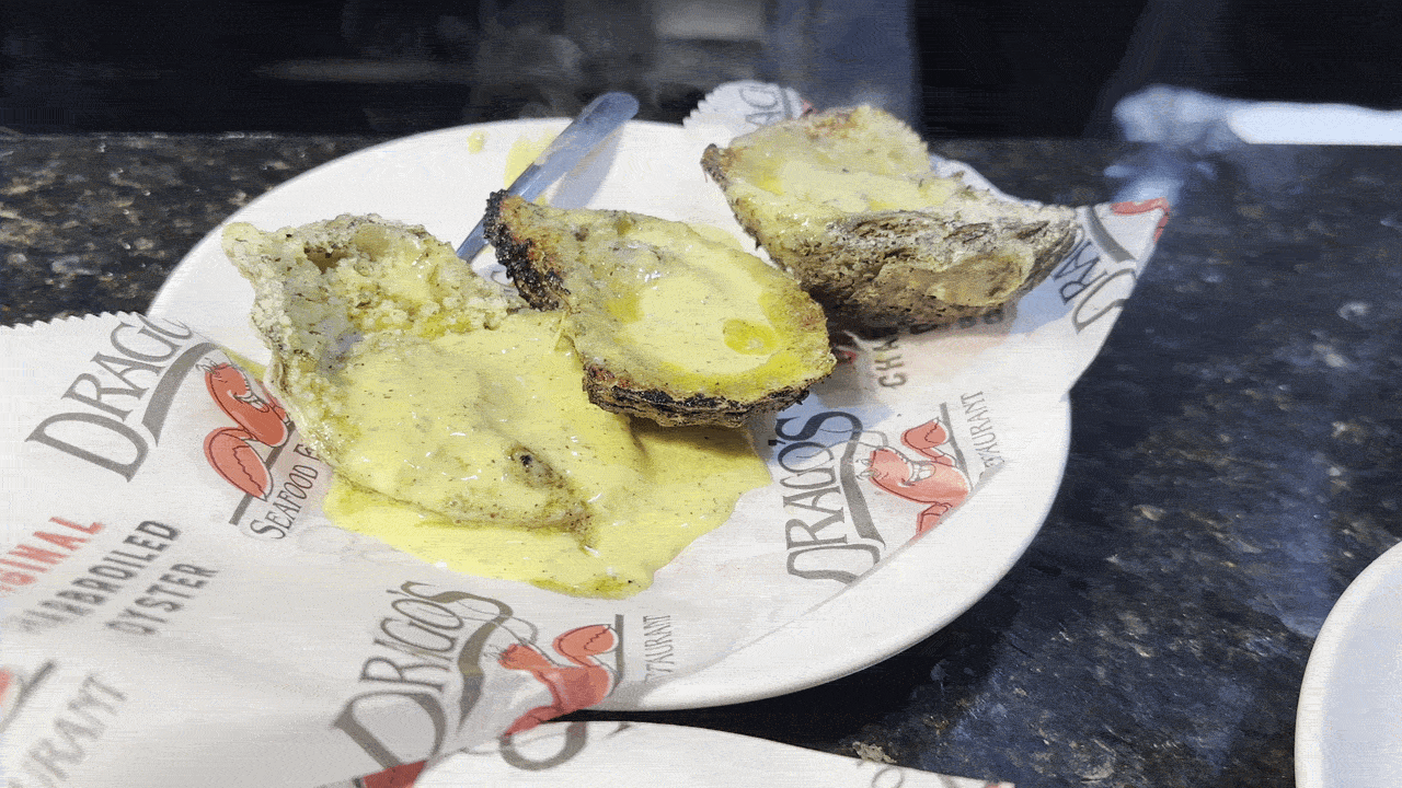 Plate with several oysters drizzled in a yellow creamy sauce. A metal utensil rests on a white plate lined with dragon-themed parchment, on a dark speckled countertop.