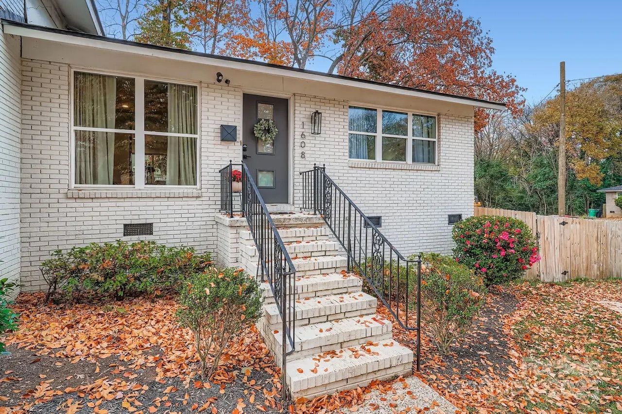 White brick house entrance with gray door adorned with wreath, black iron railings on stairs, fallen autumn leaves on ground, shrubs, and trees with fall colors in background.