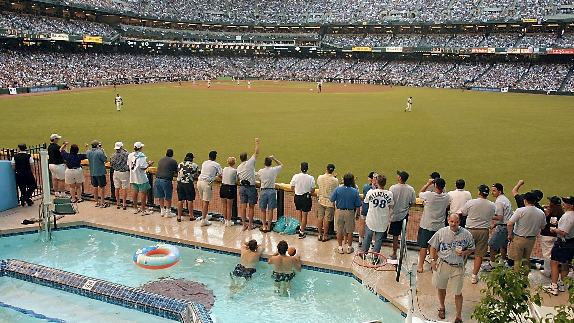Panoramic view of a packed baseball stadium, players on the diamond. In the foreground, a poolside deck with spectators leaning over the railing and two swimmers in the pool, watching the game.