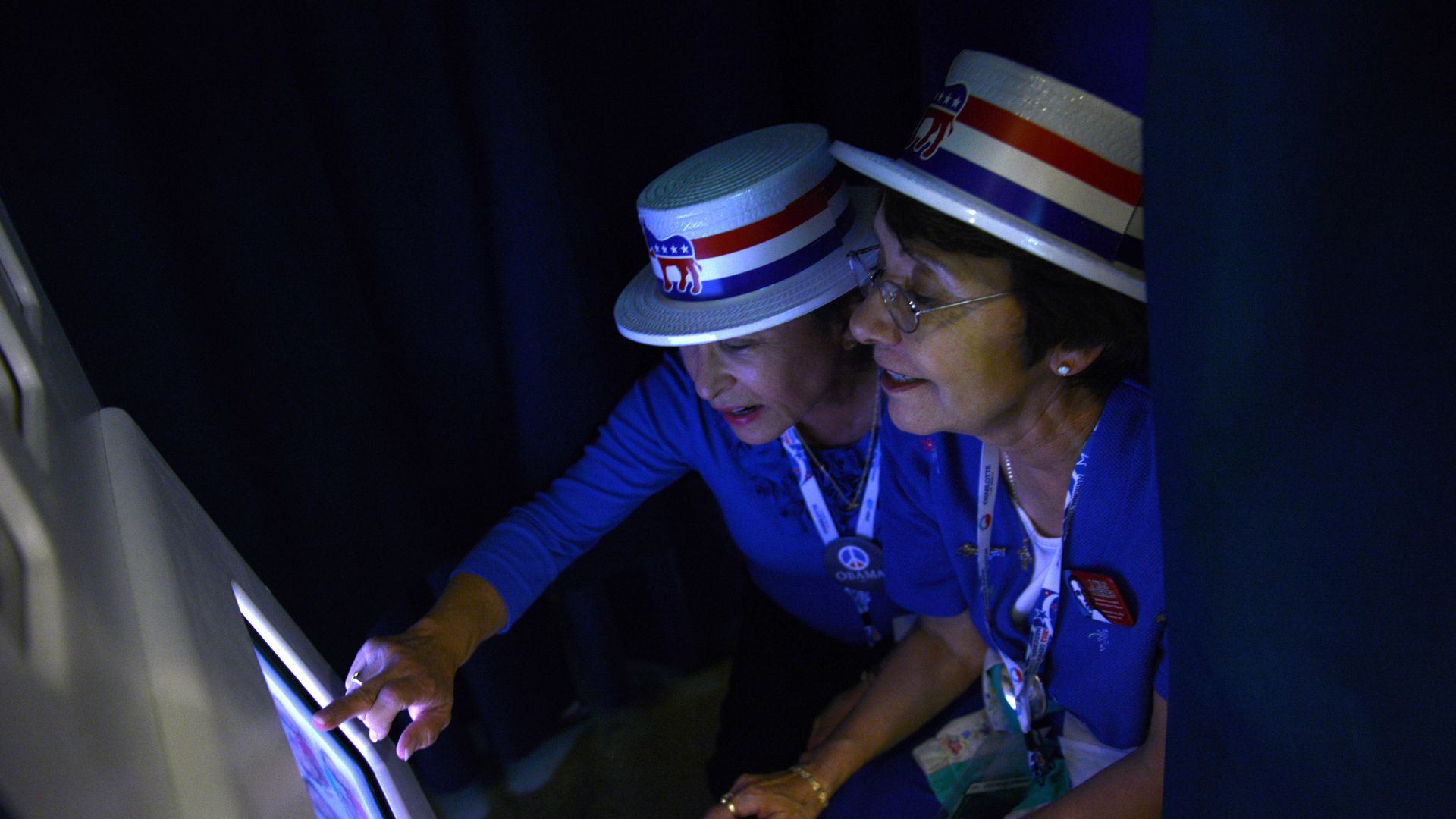 Two women in democratic red white blue hats touch a touch screen